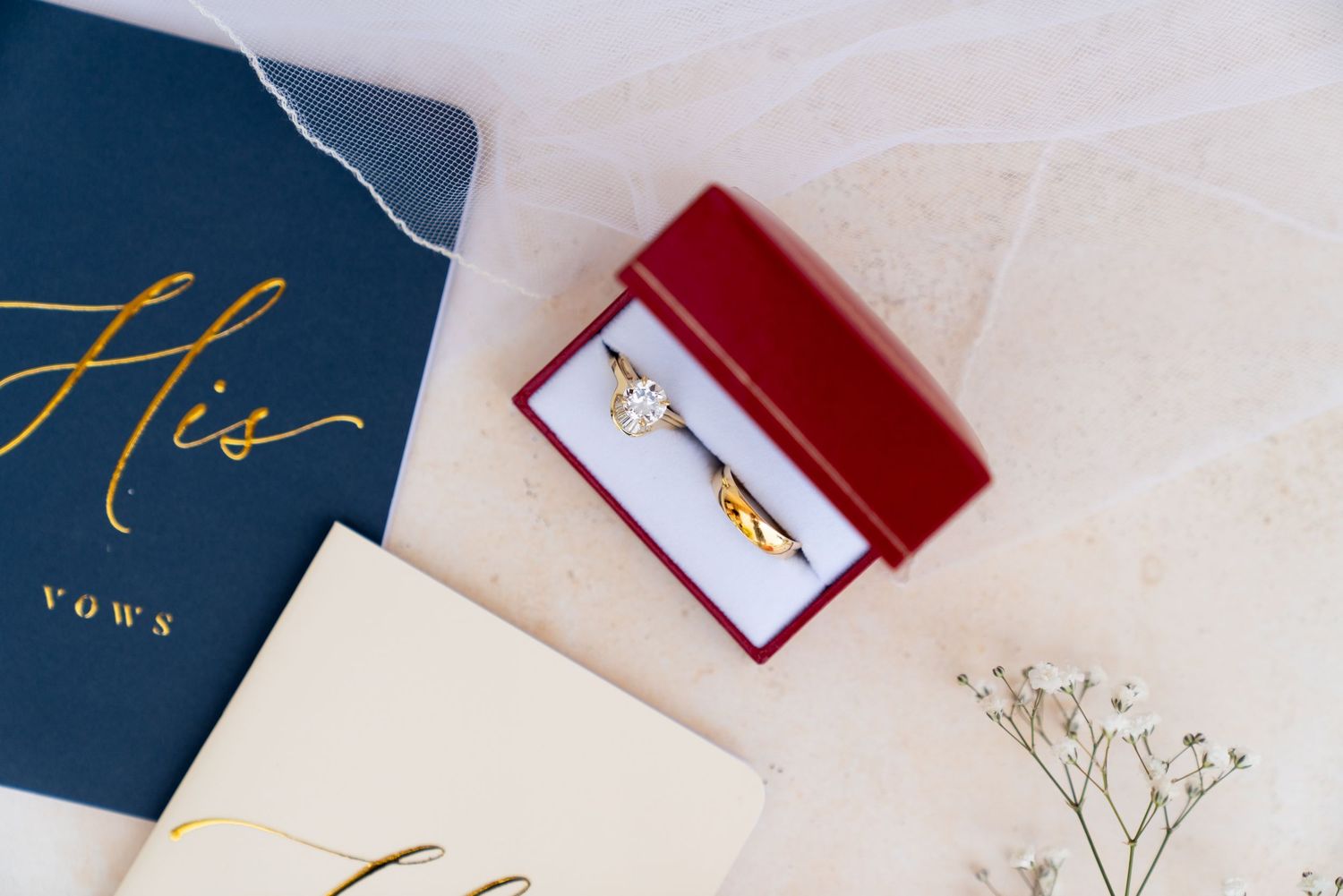 Wedding vows book in navy blue with gold text beside a red ring box and delicate baby's breath flowers.