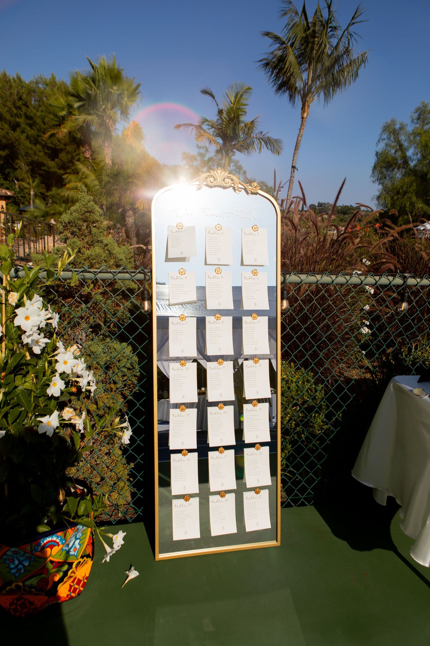 Wedding seating chart display board set up outdoors with palm trees and garden setting.