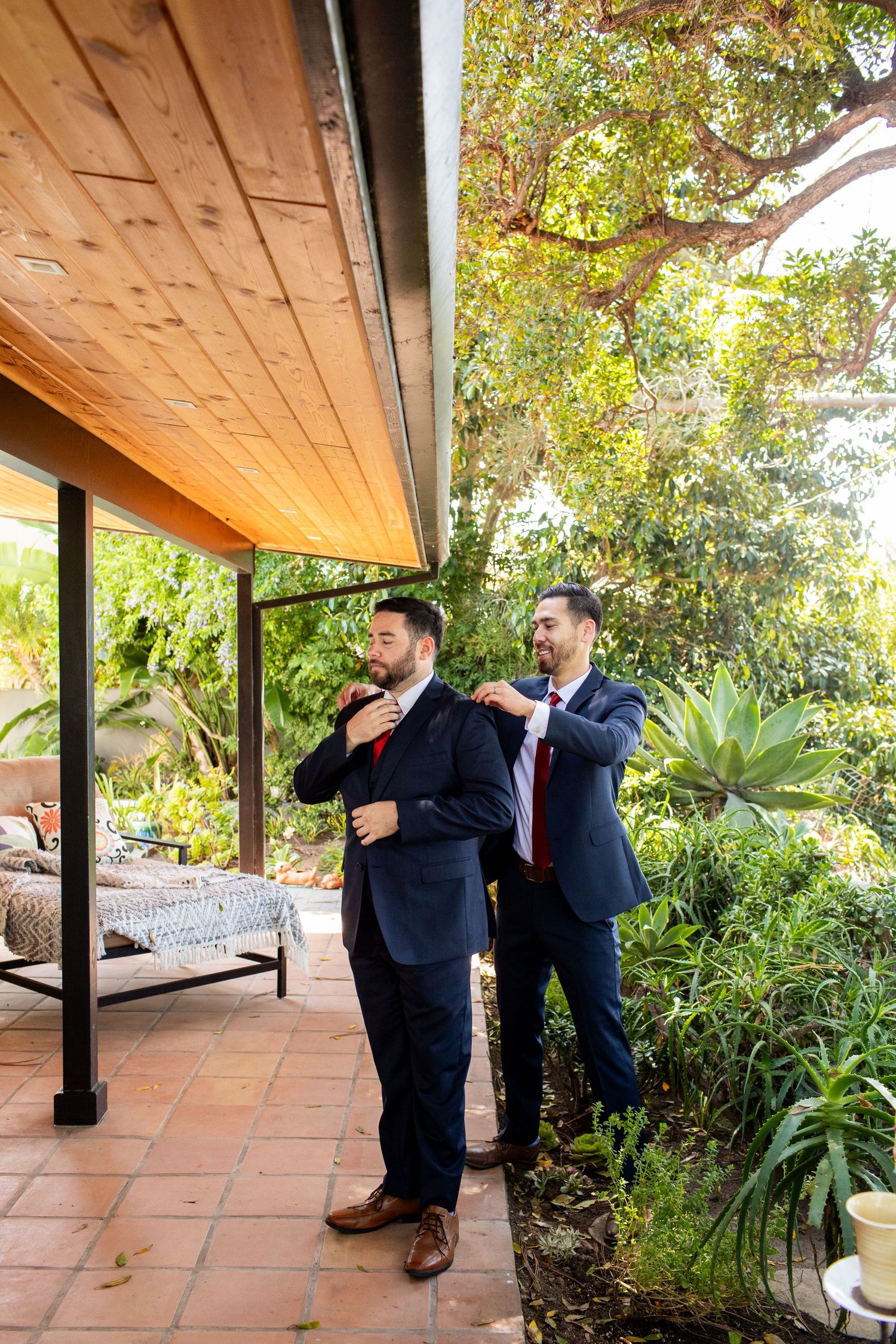 Two groomsmen sharing a lighthearted moment on a patio surrounded by lush garden greenery.