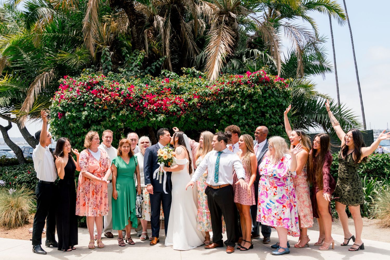 A group gathers outdoors for a celebration under palm trees and pink flowering bushes on a sunny day.