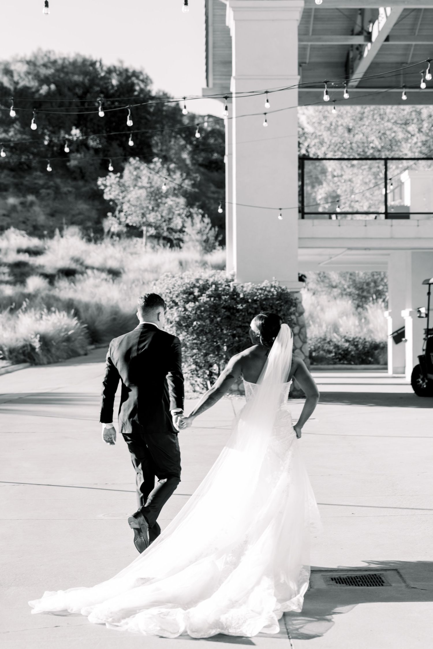 A black and white photo captures a couple walking with a flowing wedding dress trailing behind.