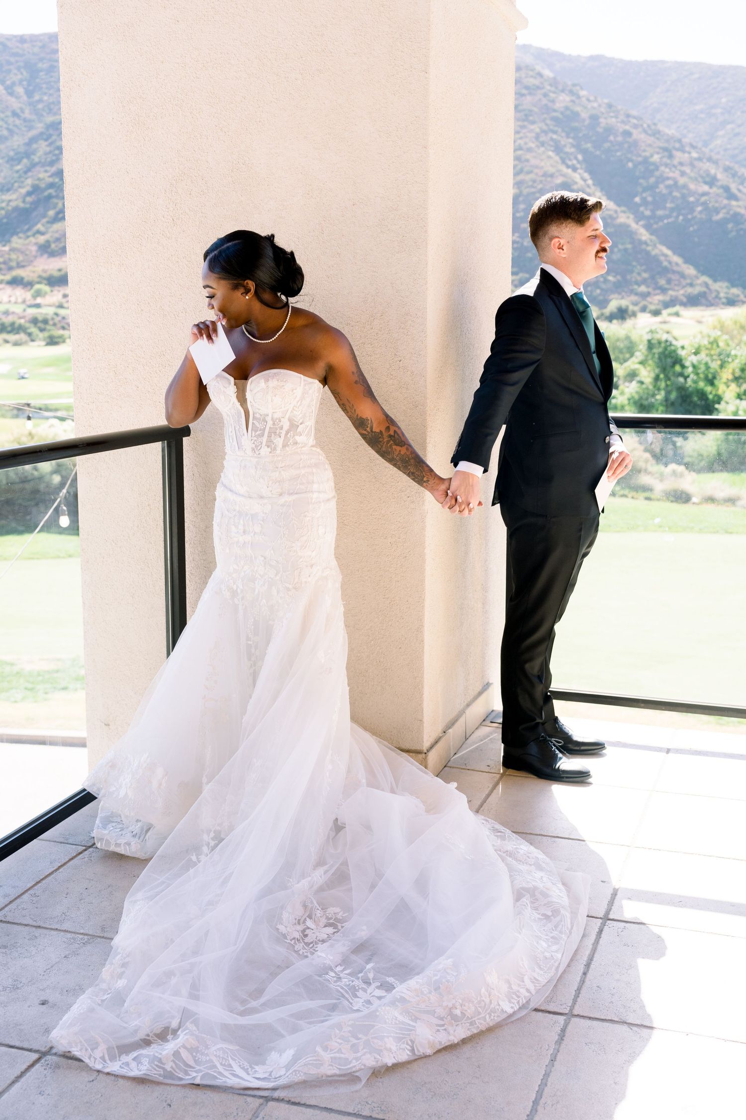 A bride in a white gown holds hands with her groom while separated by a column on a scenic balcony.