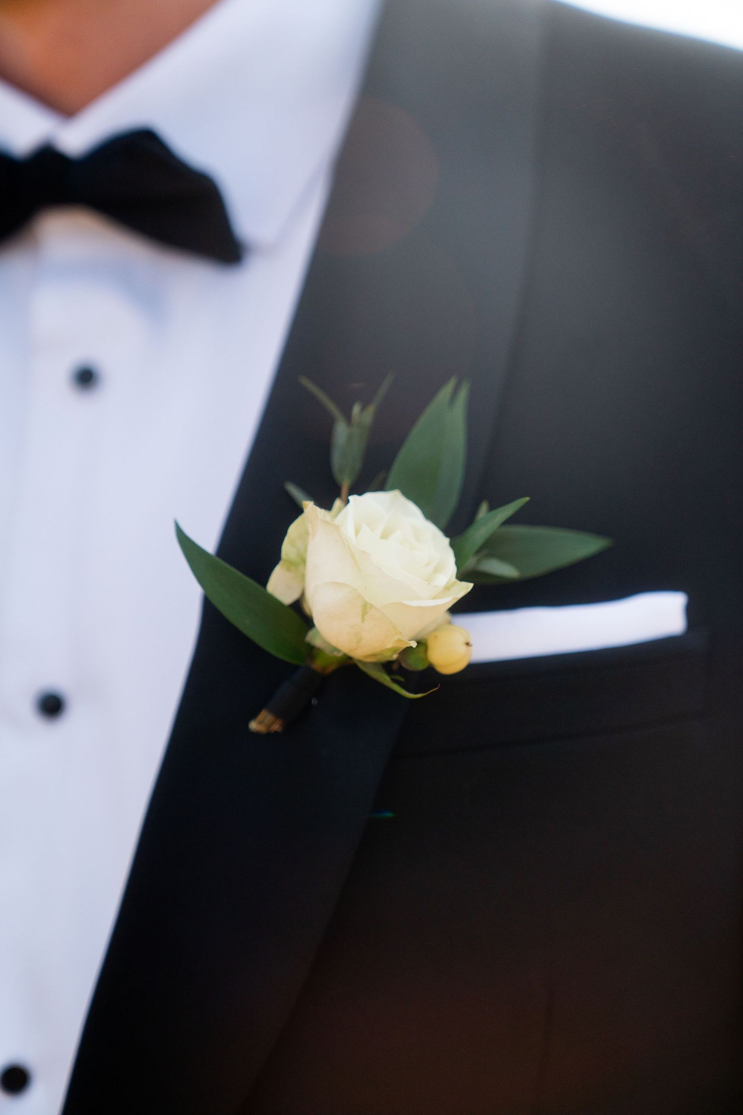 Close up of white rose boutonniere with greenery pinned to black tuxedo lapel.