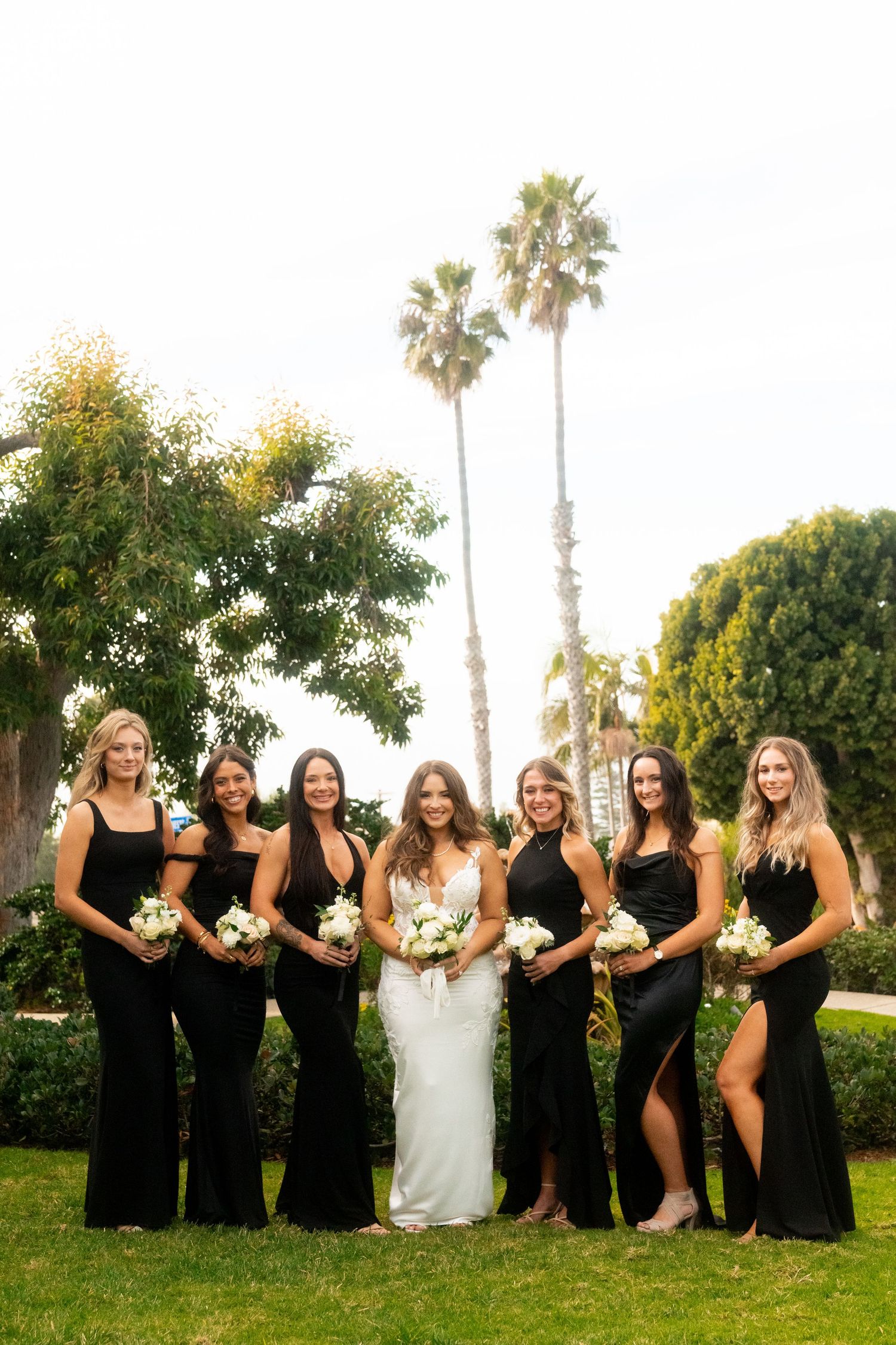 Group in black formal dresses stands with a bride in white against palm trees and lush greenery in an outdoor setting.