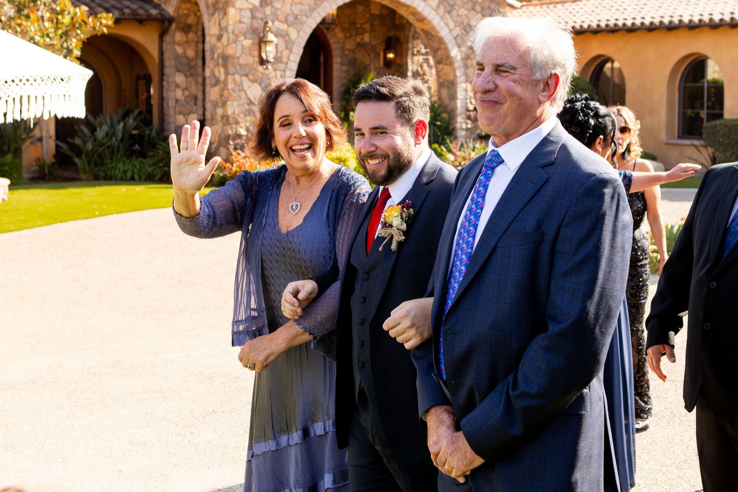 Wedding guests smile and gesture while walking together through a Spanish-style stone archway with sunny courtyard.