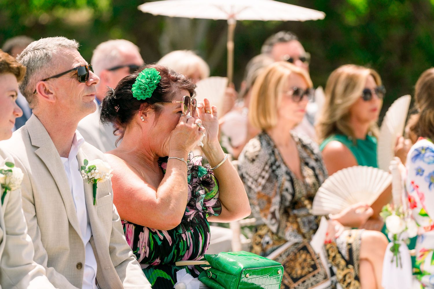 Wedding guests sitting outdoors under parasols watching ceremony in summer garden setting.