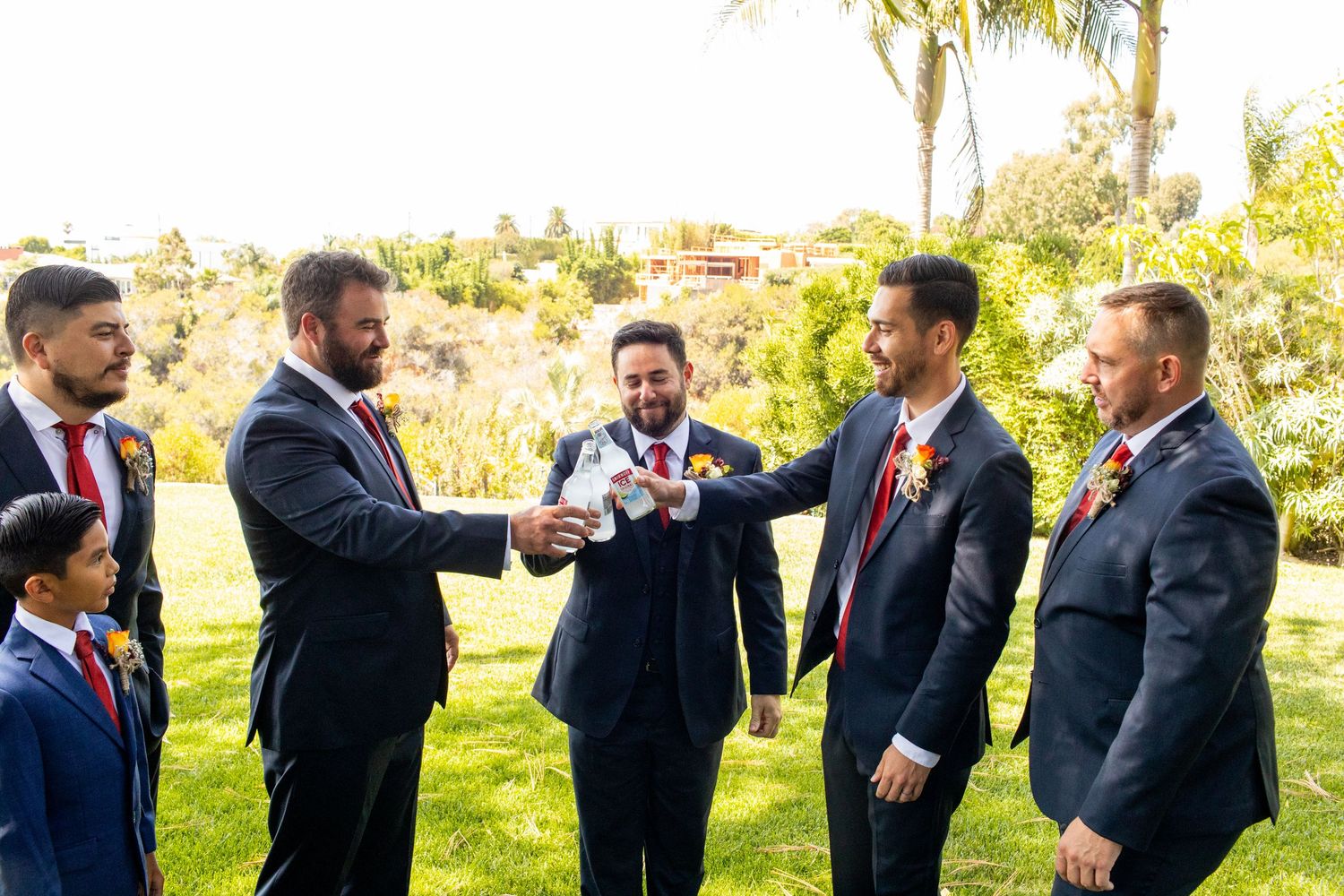 A group of groomsmen in suits toasting and laughing together outdoors on a sunny day.