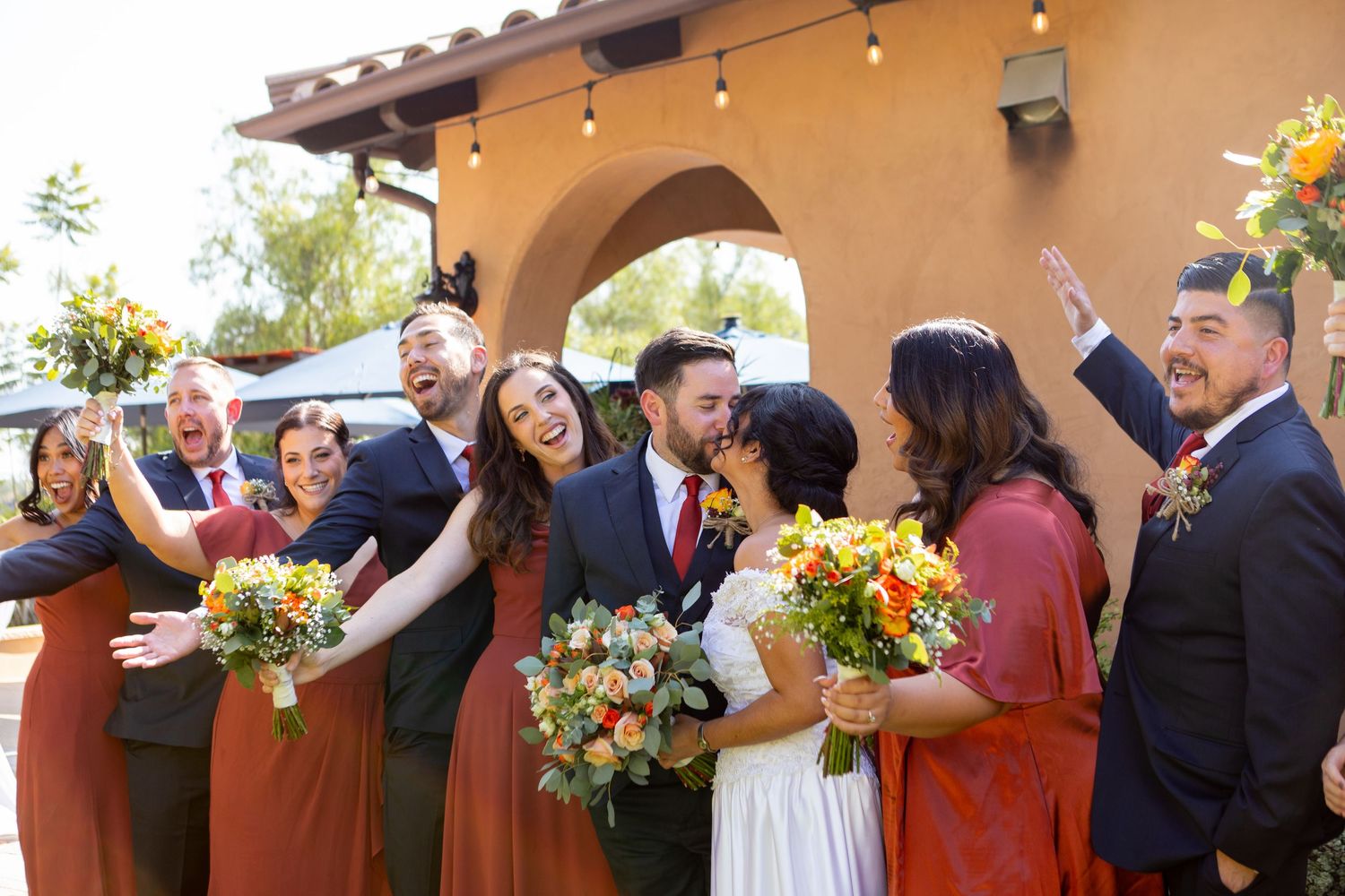 Joyful wedding party in burgundy attire celebrates under Spanish-style arches with bright sunlight and flowers.