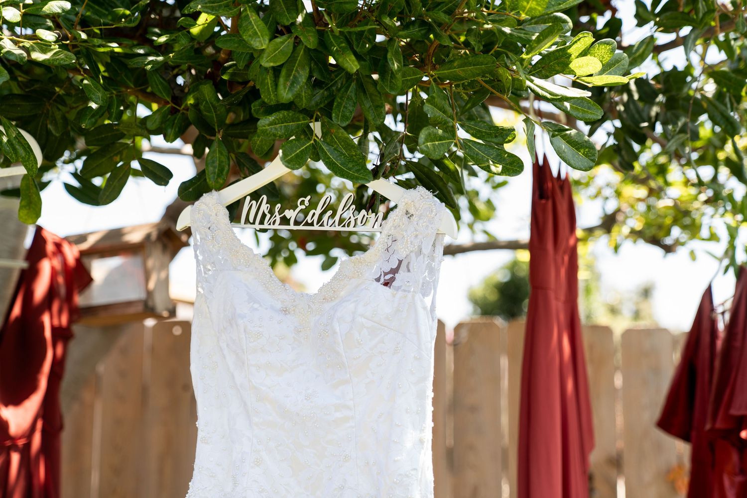 A white lace wedding dress hangs from a decorative hanger under green tree branches against a blurred background.