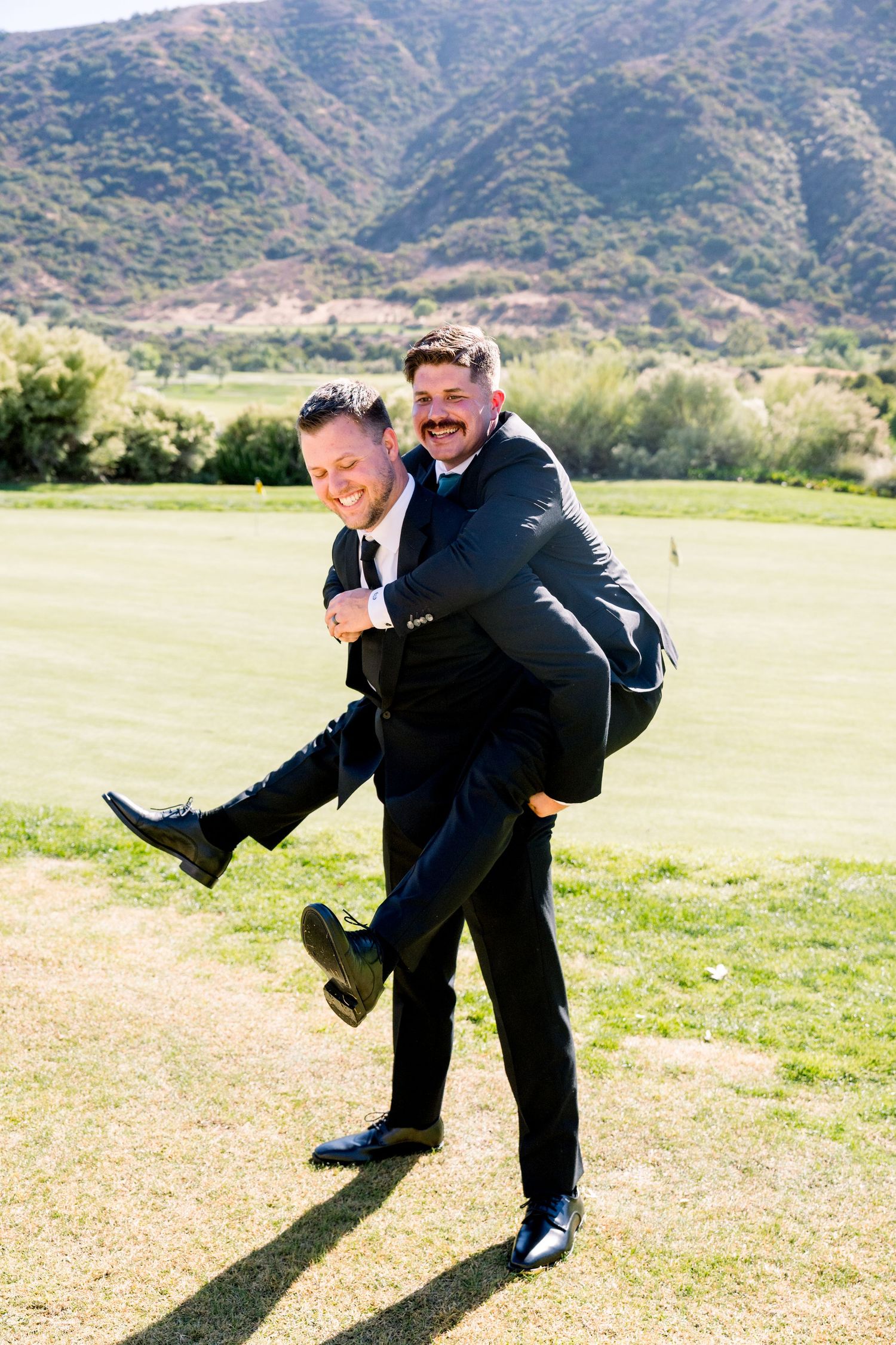 Two people in suits playfully posing on a grassy field with mountains in the background.