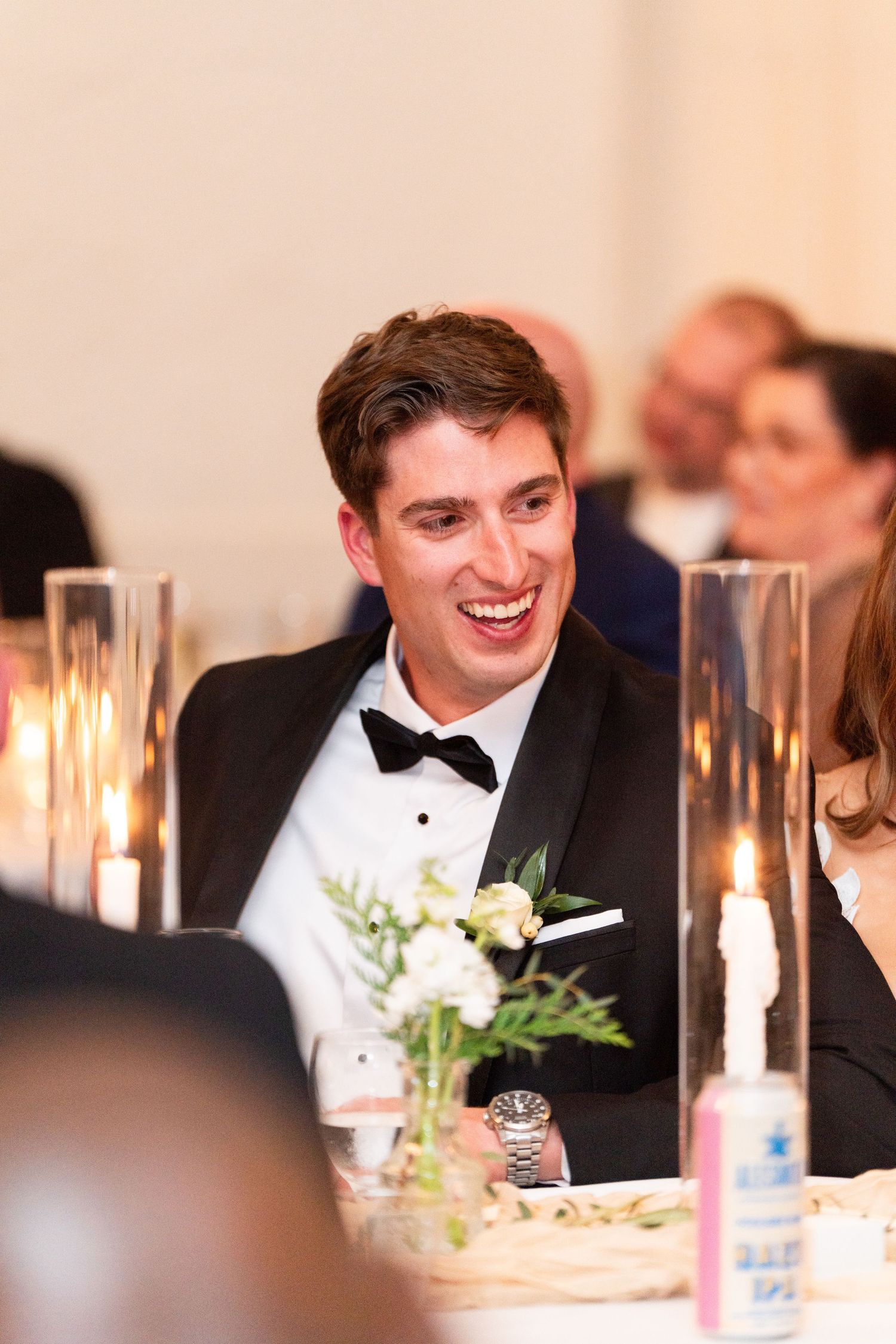 A person in a black bow tie and tuxedo smiles while seated at an elegant candlelit reception dinner table.