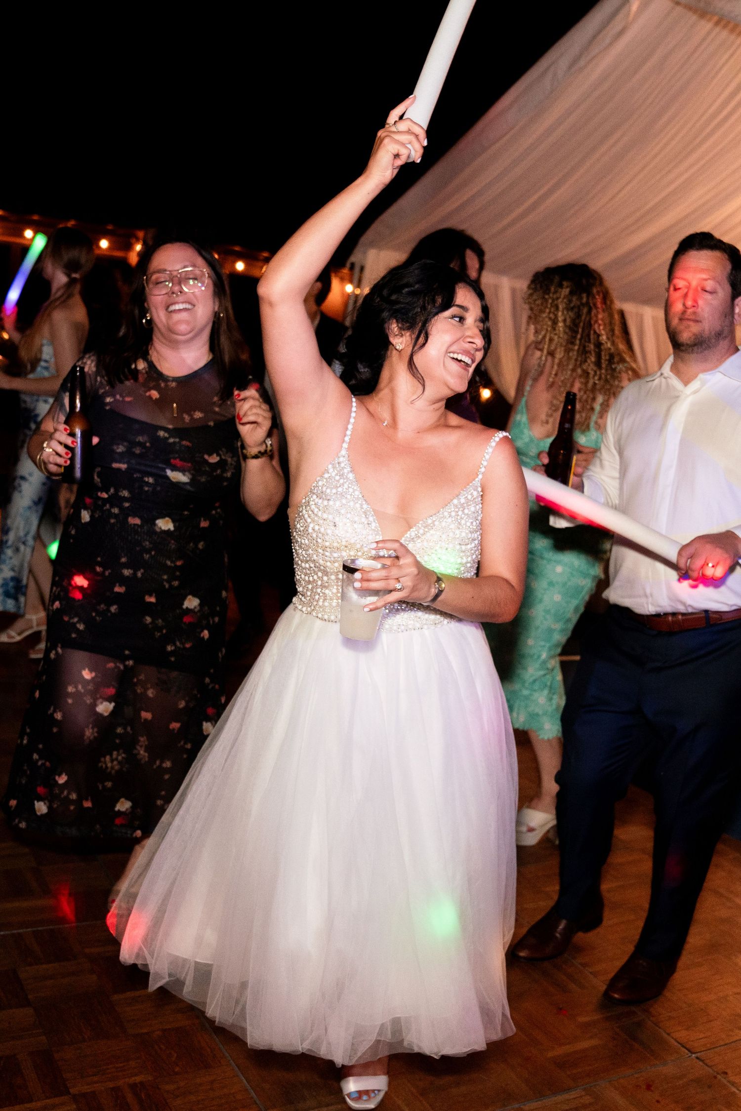 Festive wedding reception scene with dancing under colorful lights and glowsticks in a tented venue.