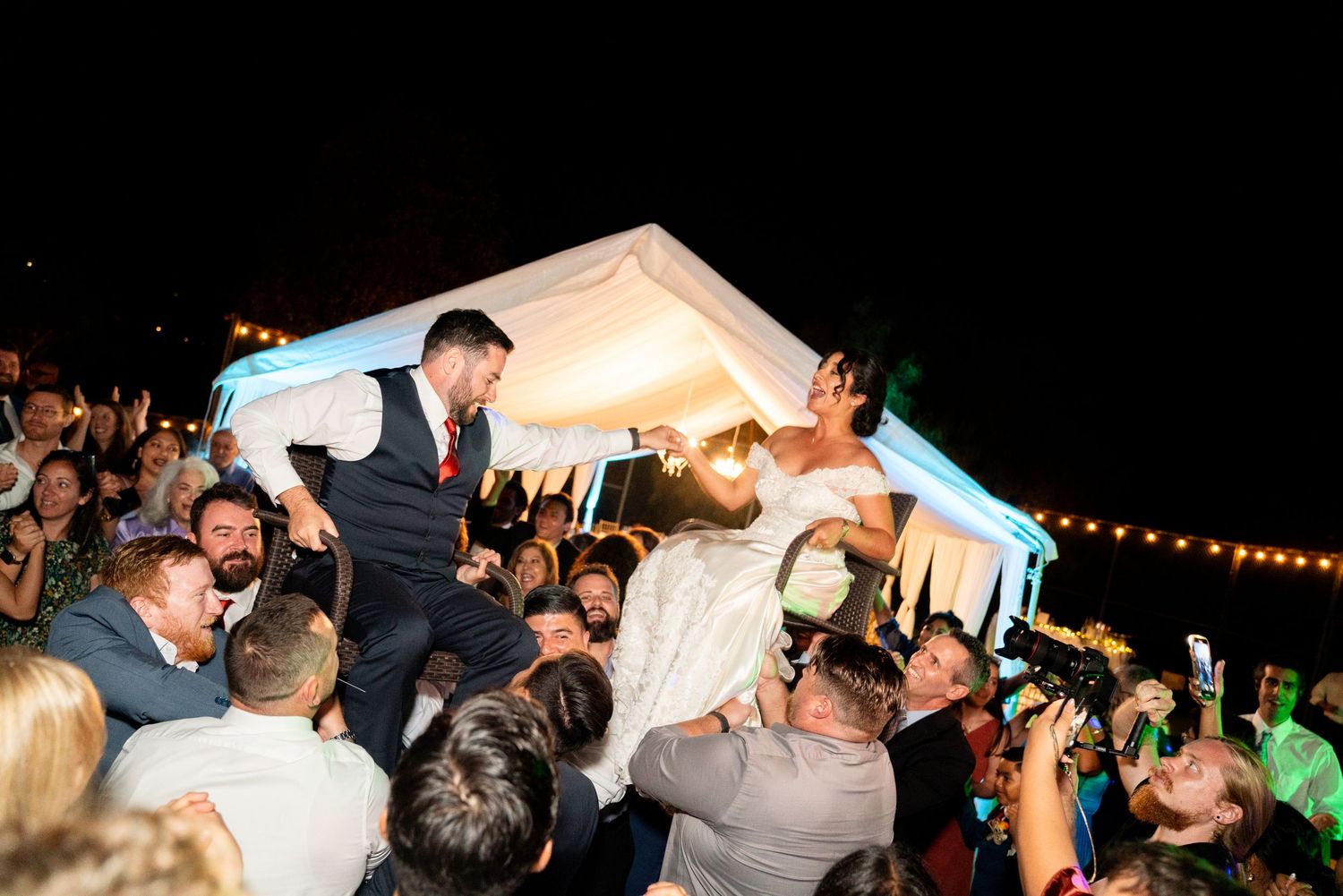 Night reception celebration with guests lifting bride and groom on dance floor.
