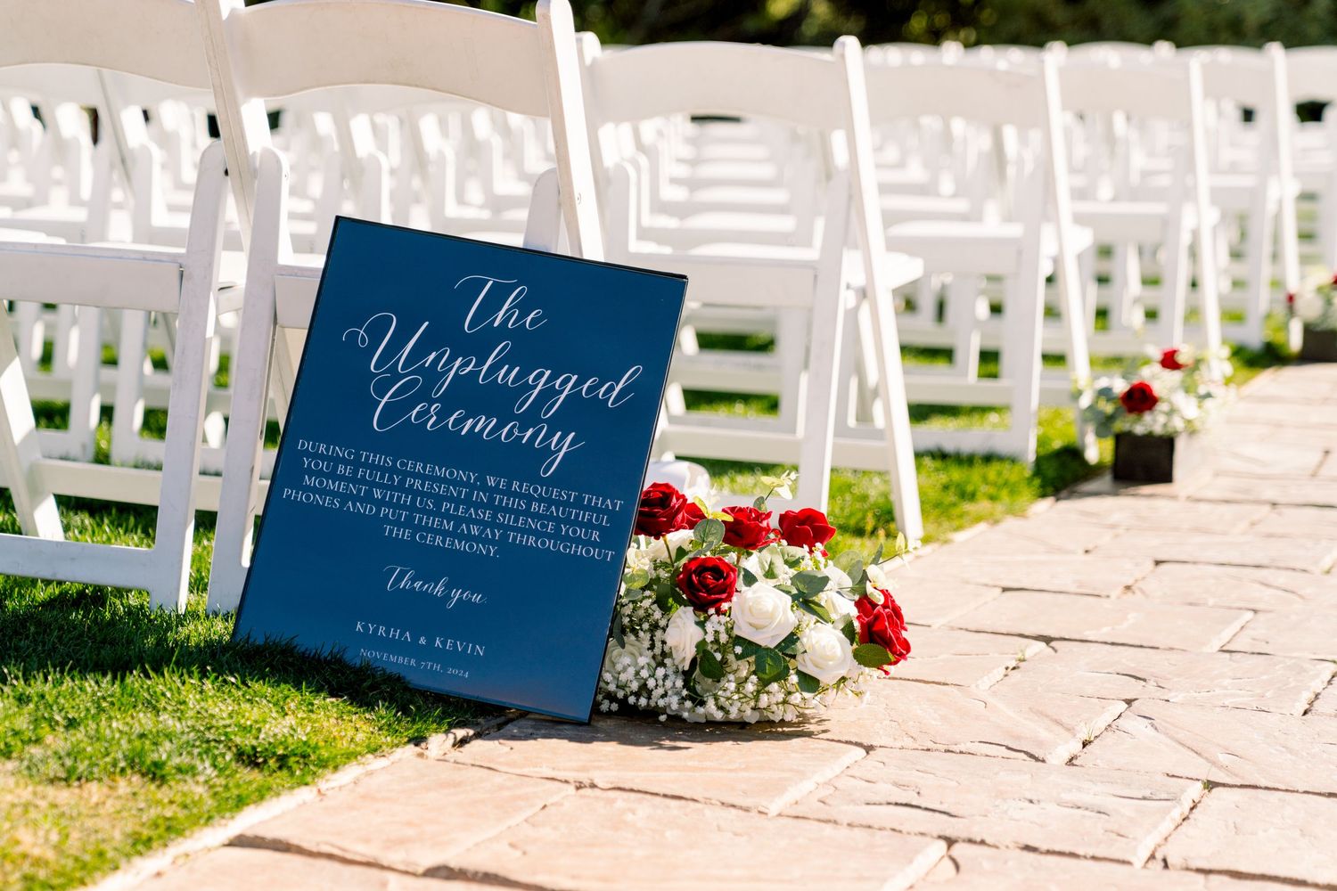 A blue wedding welcome sign sits beside white chairs and red flowers on a stone pathway.