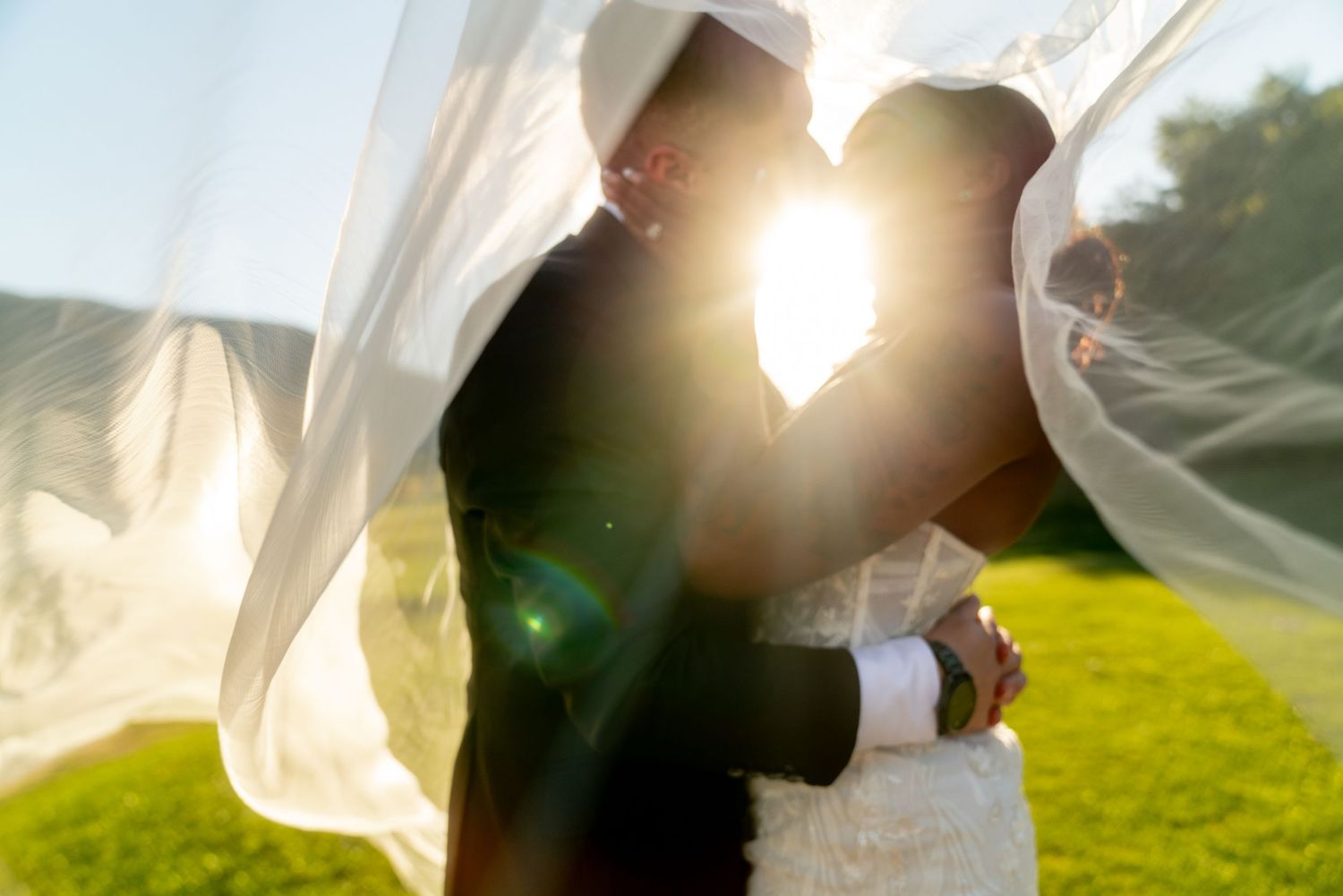 Silhouette of couple embracing under flowing veil with golden sunlight behind them.
