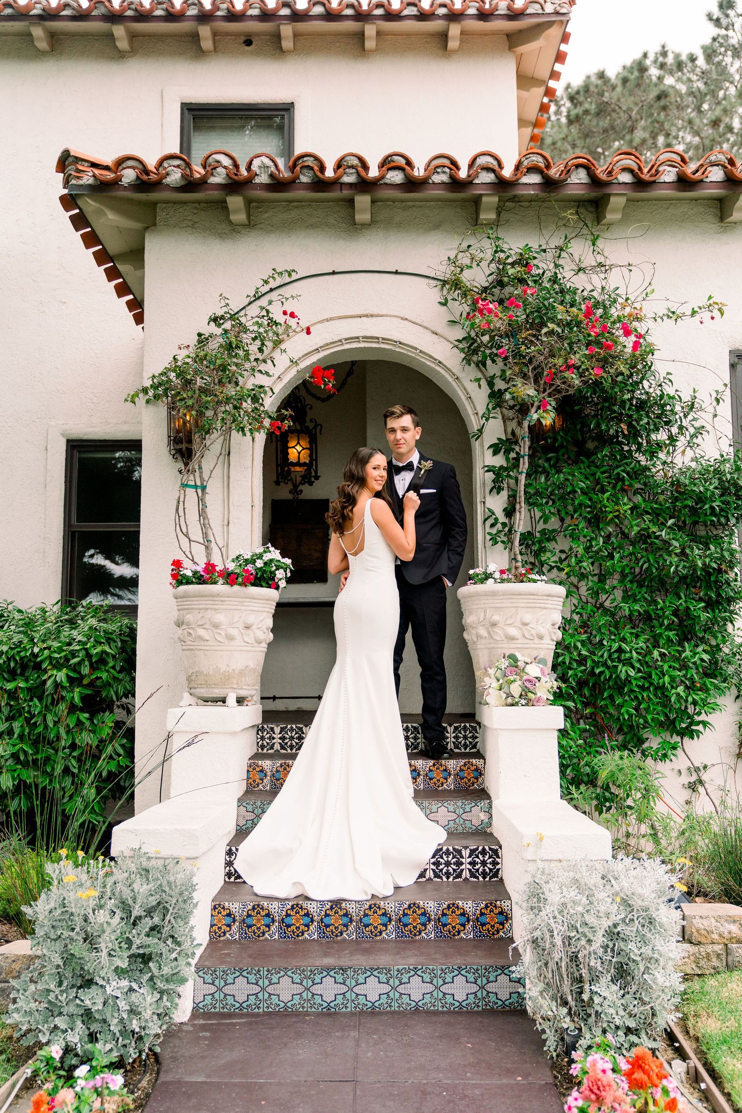Elegant wedding couple posing on Spanish-style porch with tiled steps and flowering vines.