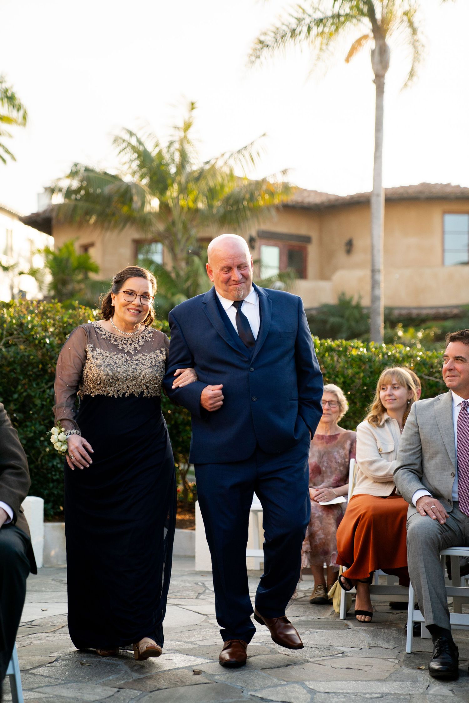 A wedding processional walking down a stone path surrounded by palm trees and guests.