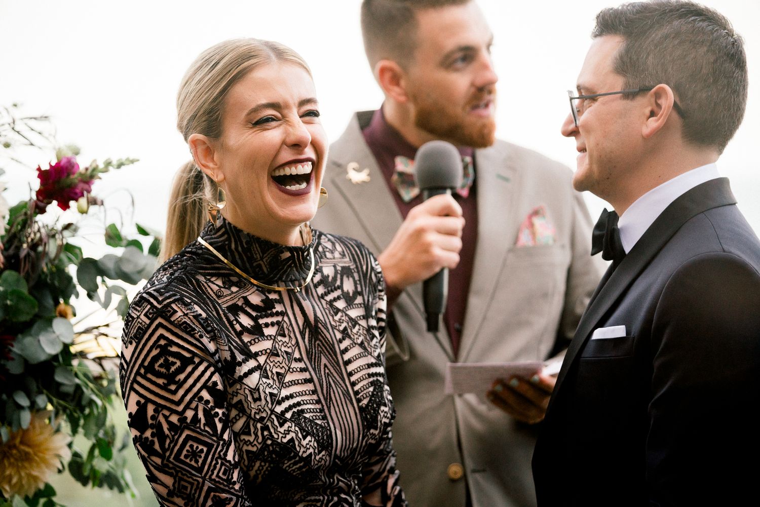 Wedding guests share a joyful moment during an outdoor ceremony with floral decor in the background.