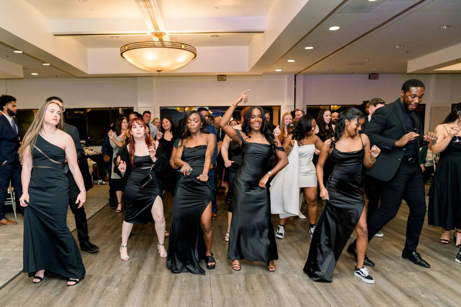Wedding party in coordinated black attire dancing in formation on reception dance floor.