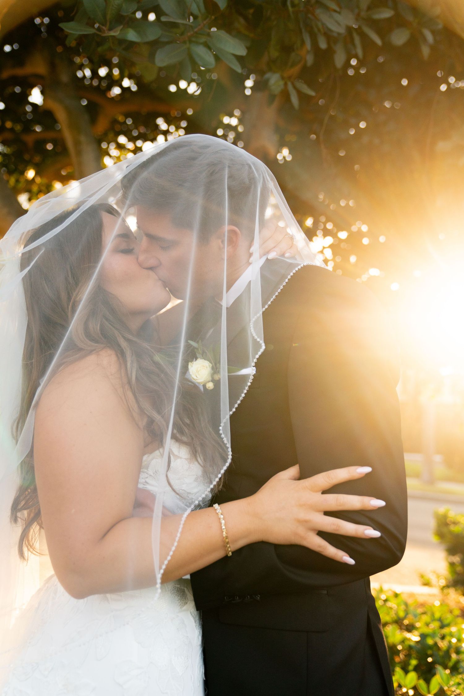Intimate wedding moment captured under bride's veil with golden sunset light streaming through trees.
