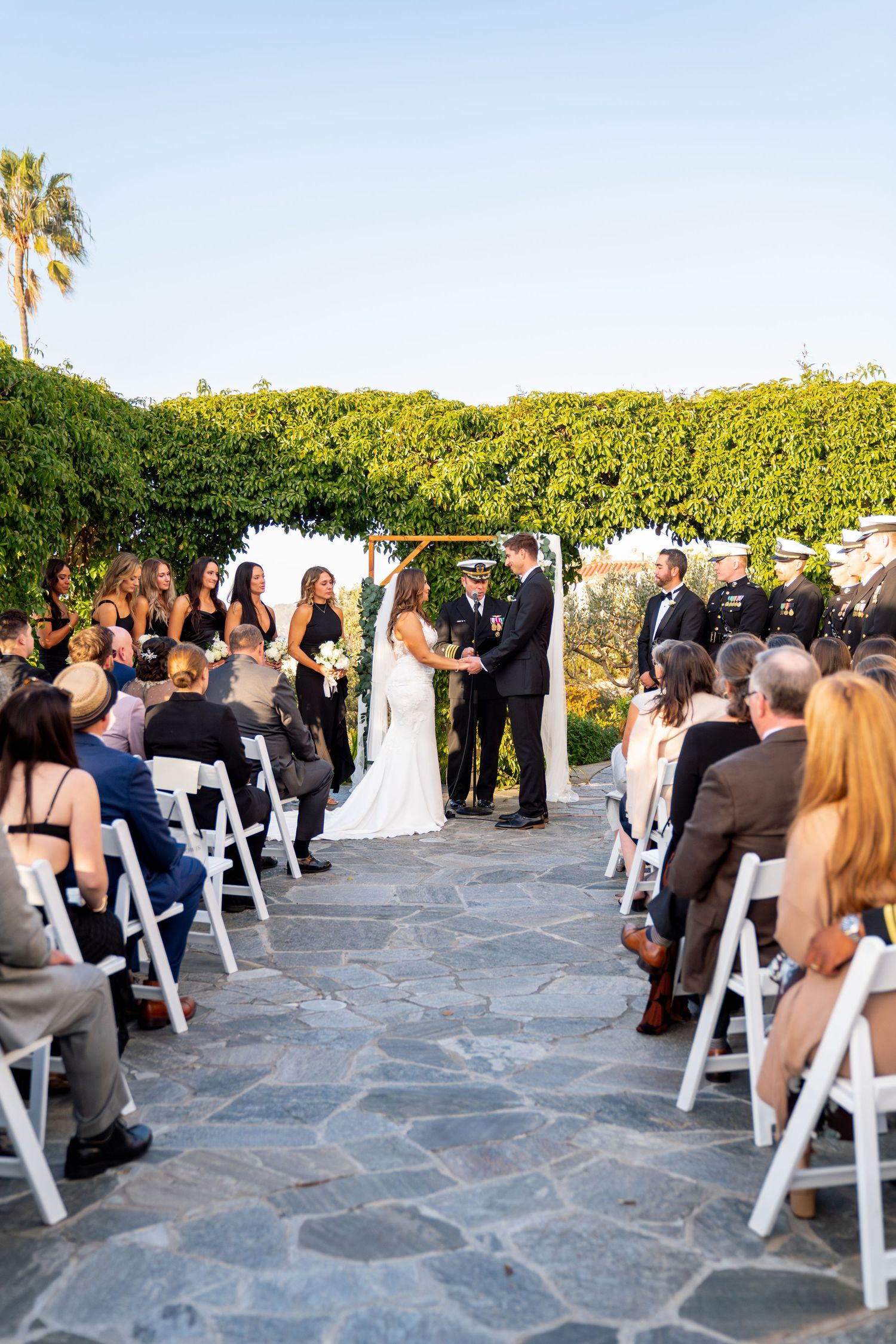 Outdoor wedding ceremony with guests seated in white chairs on stone patio under greenery.