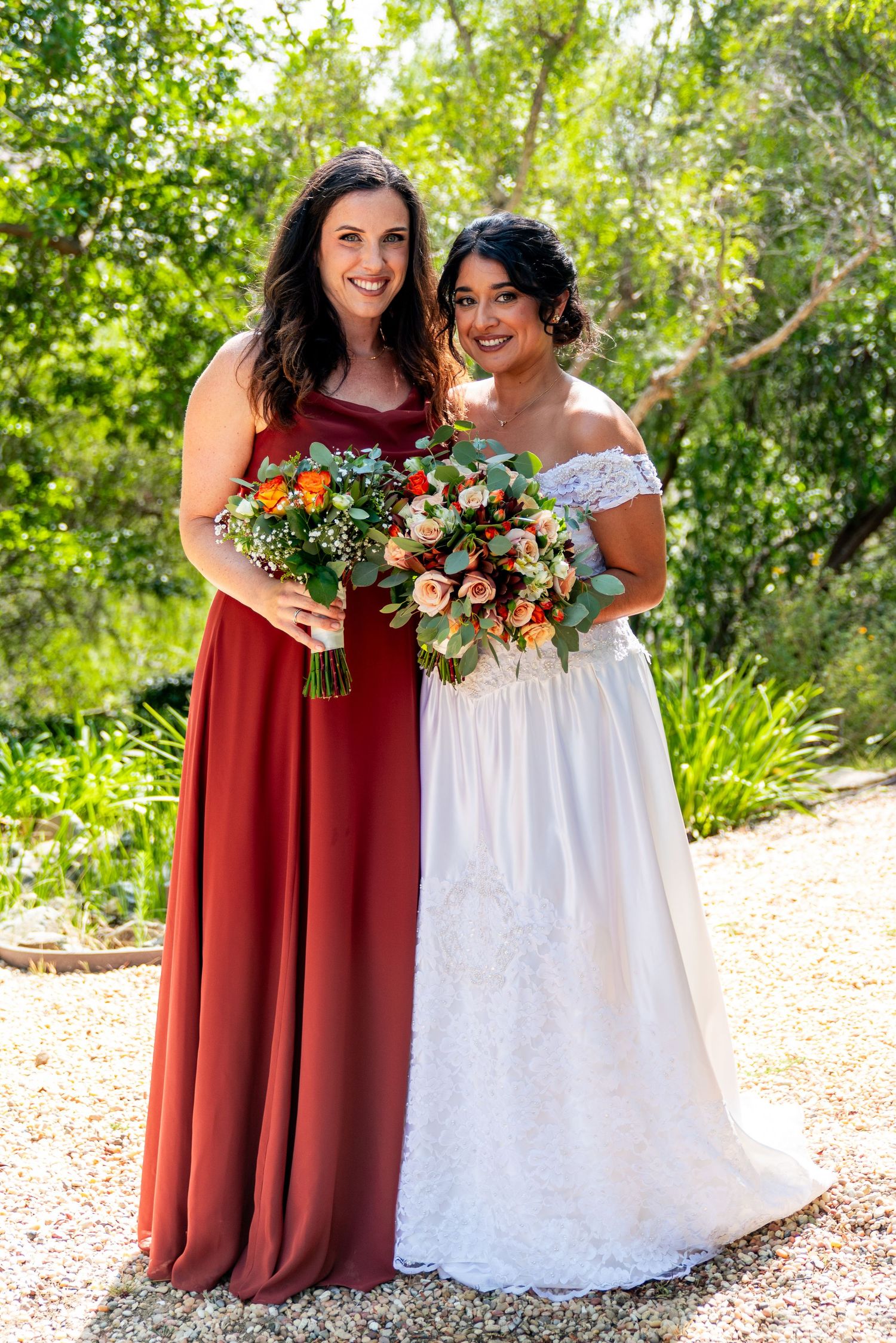 A bridesmaid in burgundy and bride in white holding matching floral bouquets pose together in a garden setting.