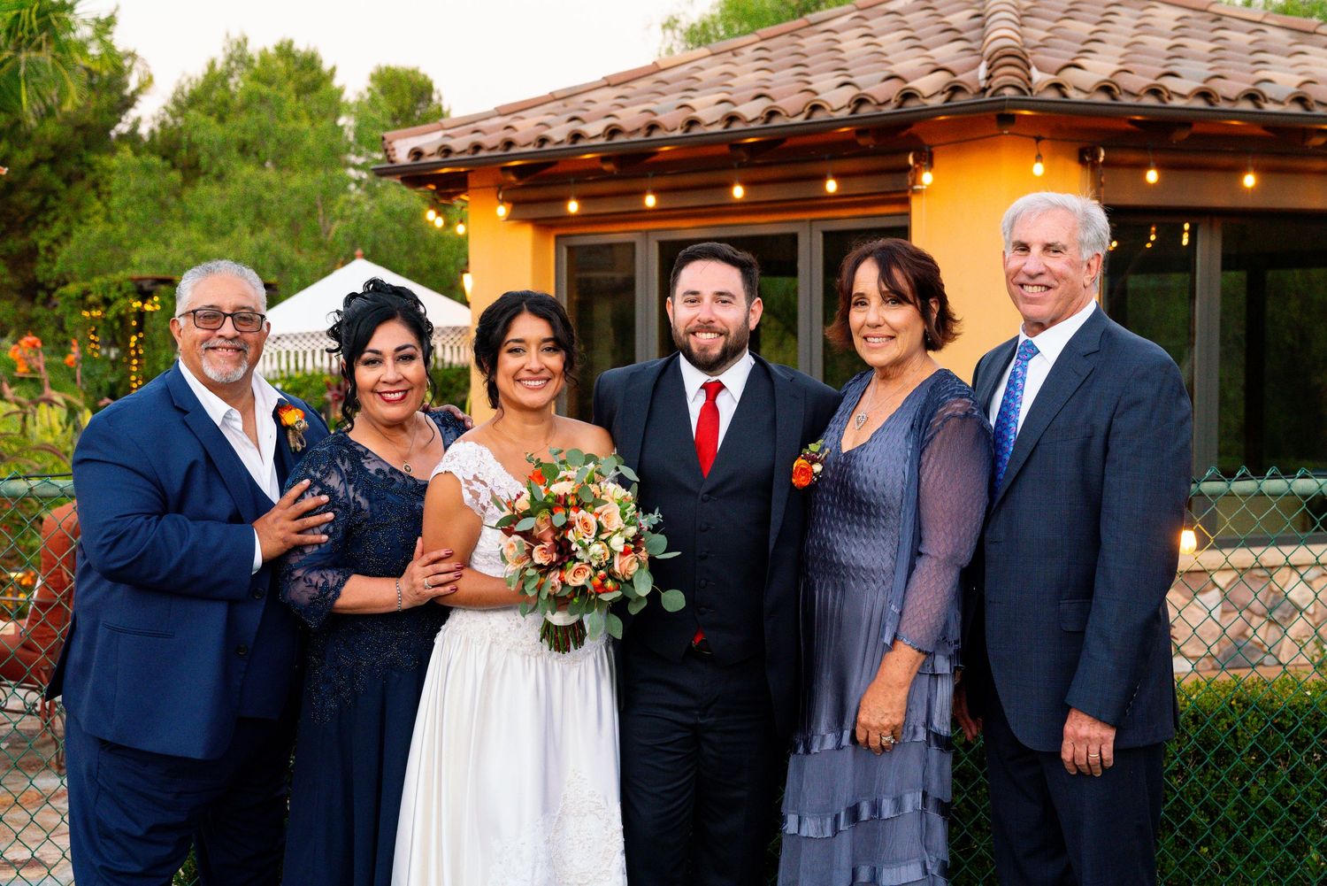 Wedding party poses for a formal photo outside a Spanish-style venue with string lights.