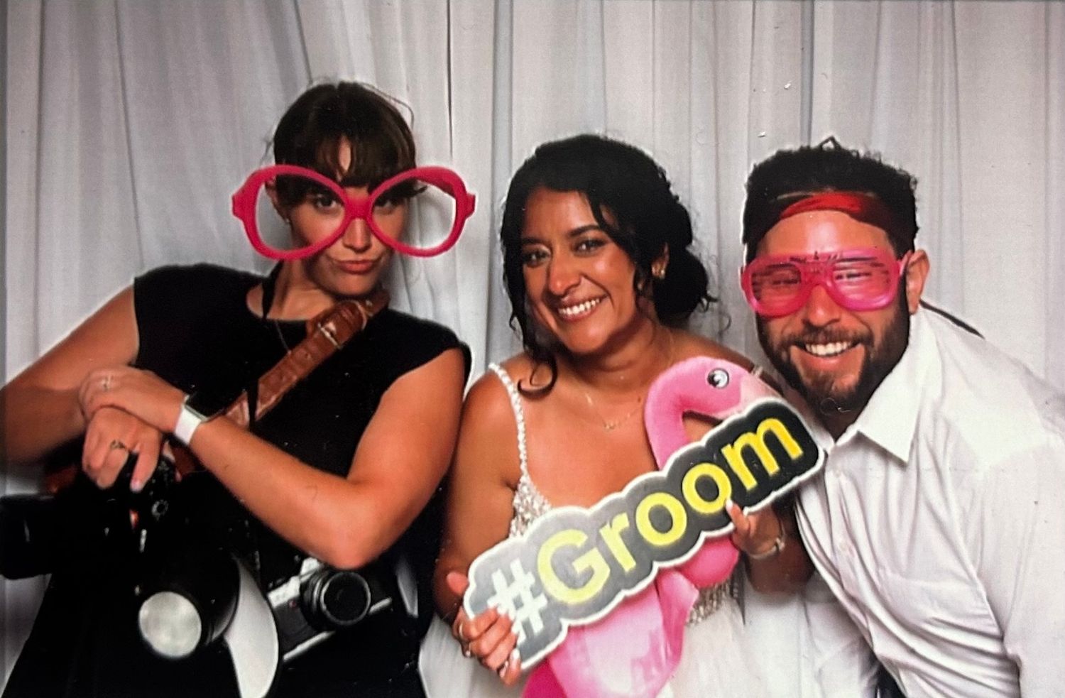 Three people posing together in a photo booth with fun pink accessories and groom signage for a wedding celebration.