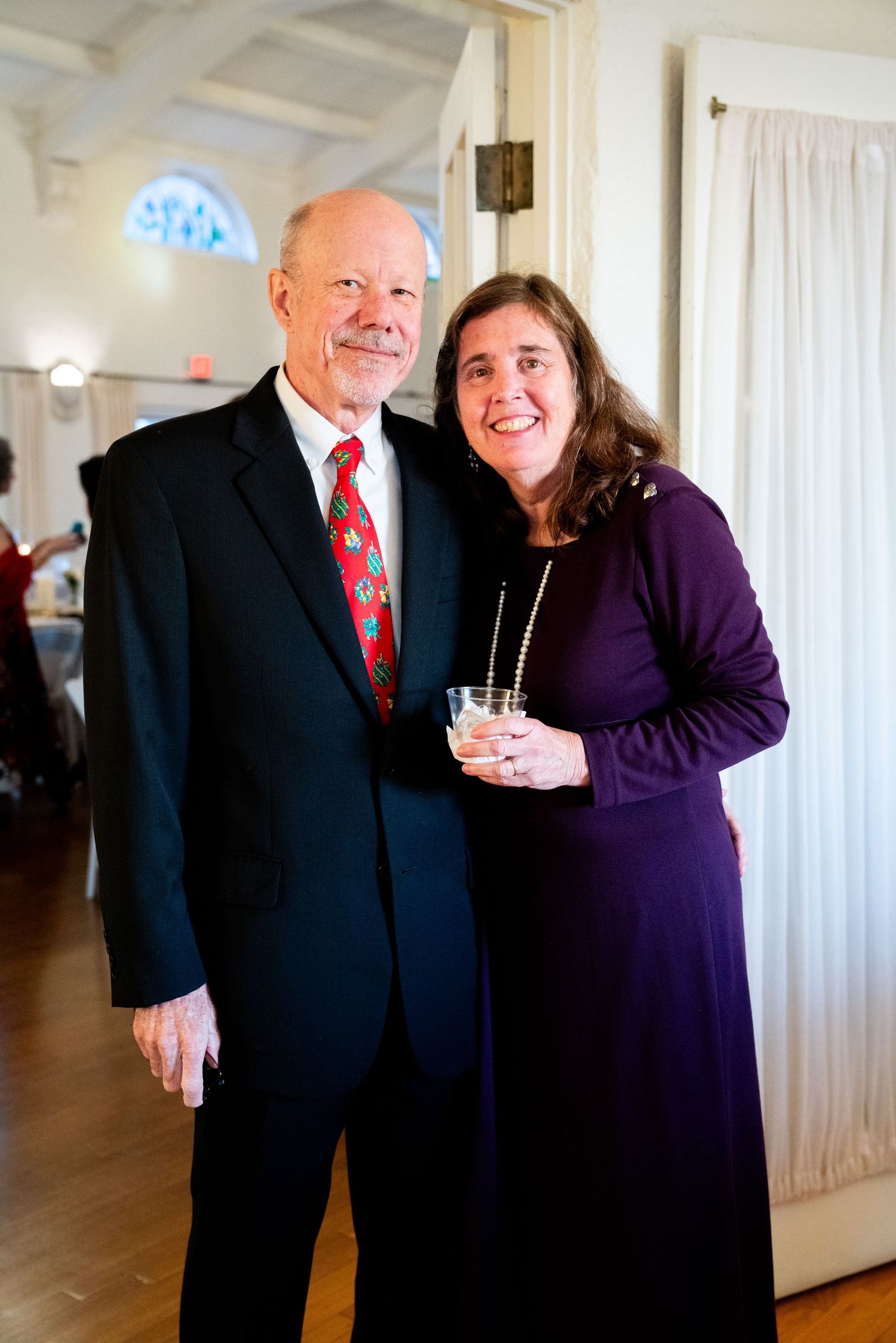 A couple poses together at a formal event, with the woman holding a drink.