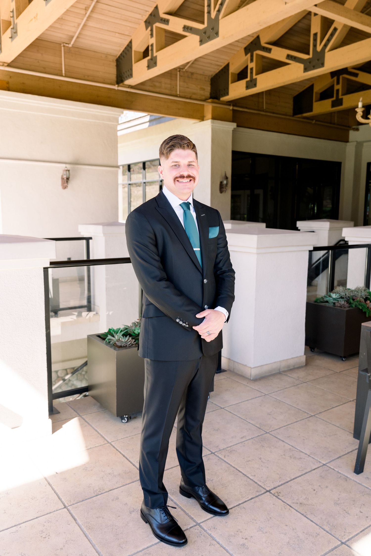 A formal portrait of a person in a dark suit and blue tie standing on a tiled terrace.