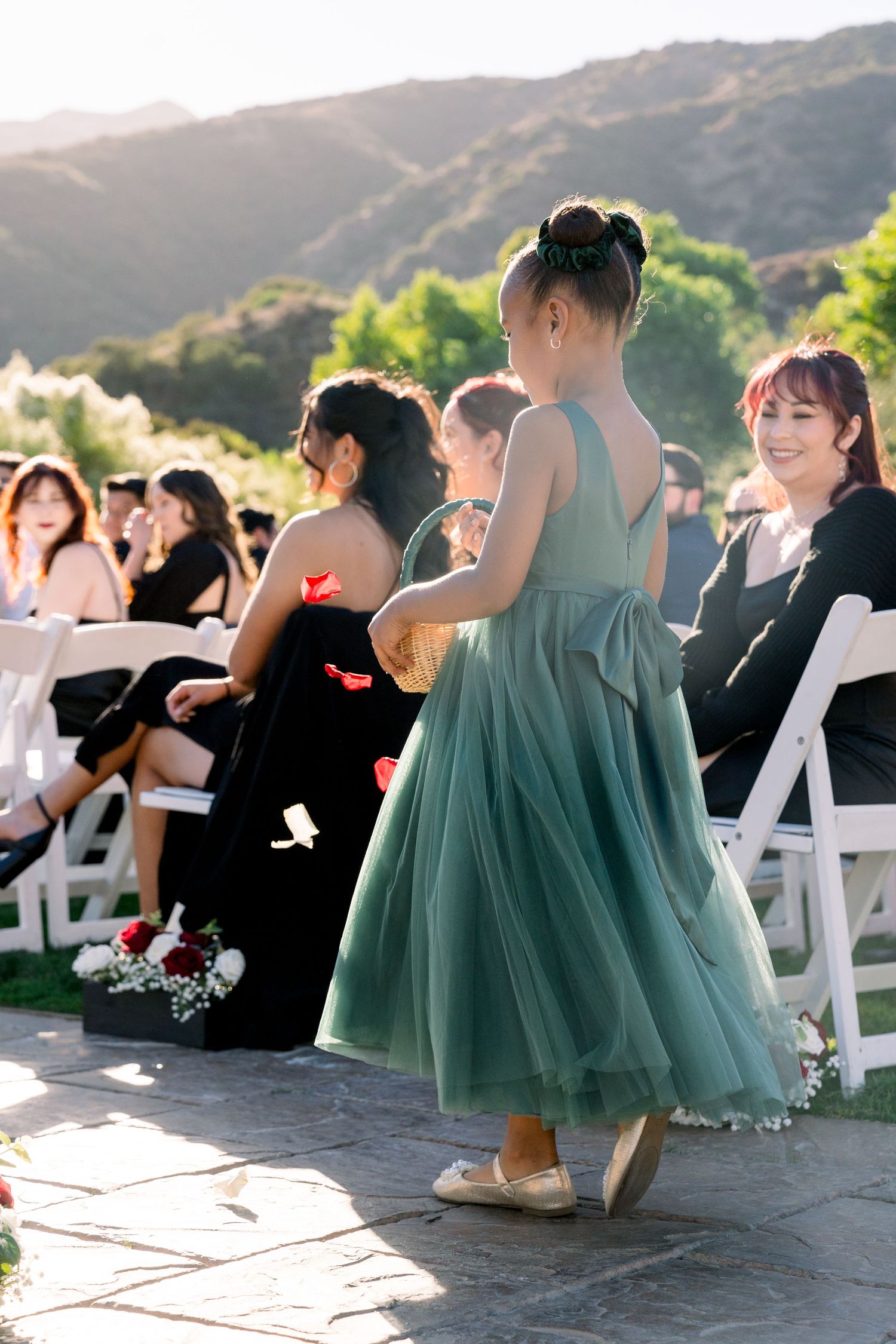 A young attendant in a mint green tulle dress stands during an outdoor wedding ceremony as guests sit in white chairs.