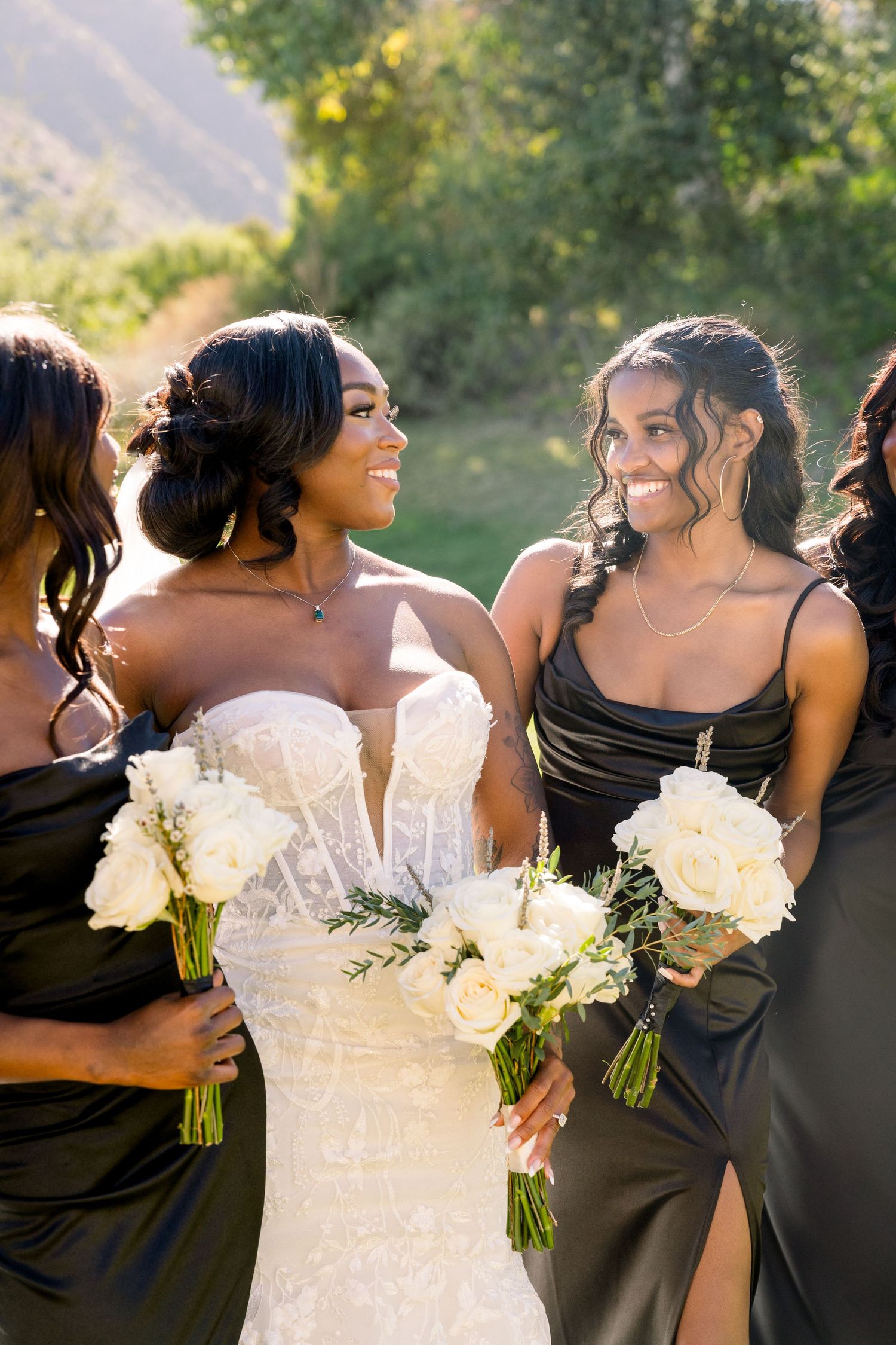 Bride in white dress shares joyful moment with bridesmaids in black dresses holding white bouquets.