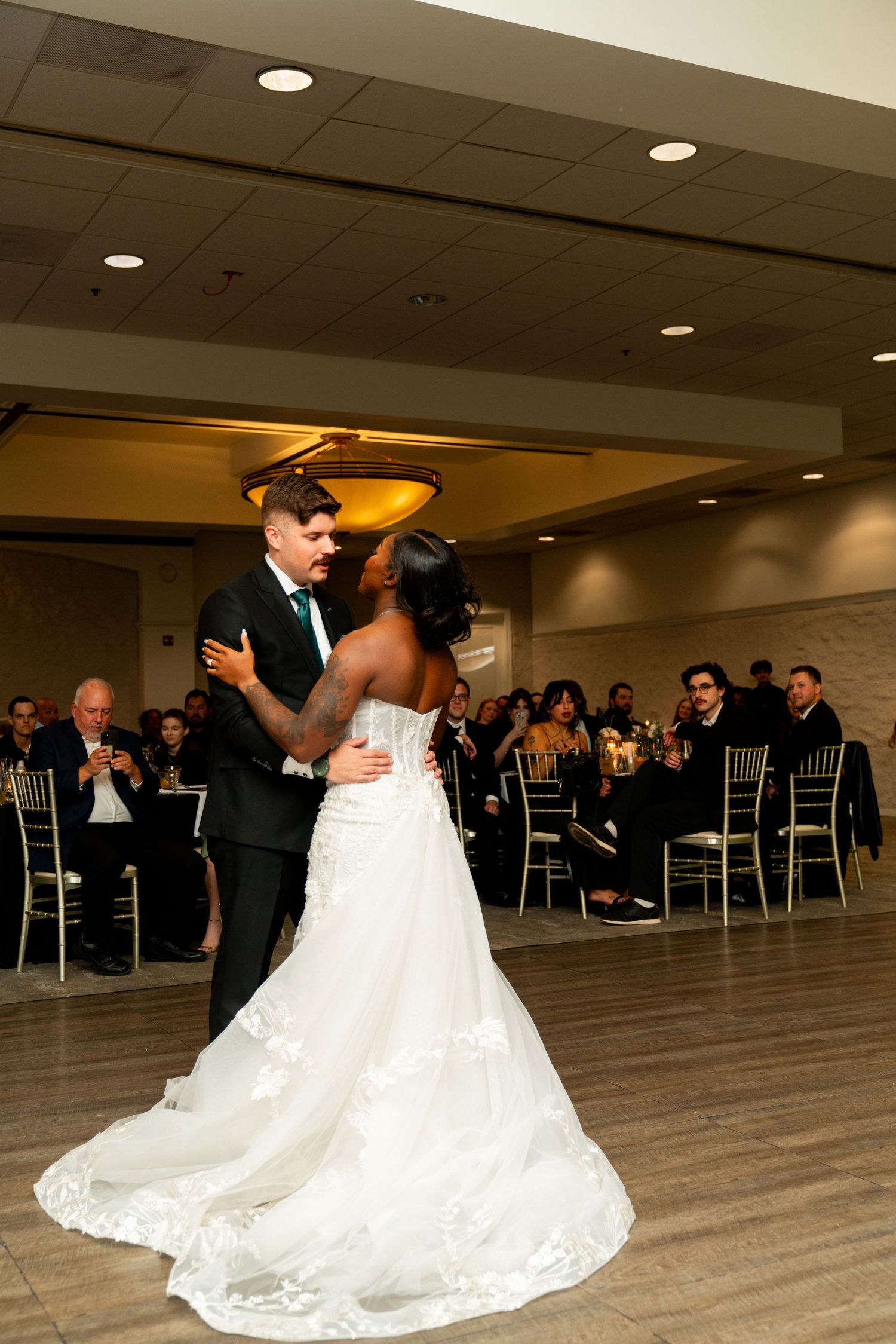 A couple shares their first dance at their wedding reception with guests seated at tables in the background.