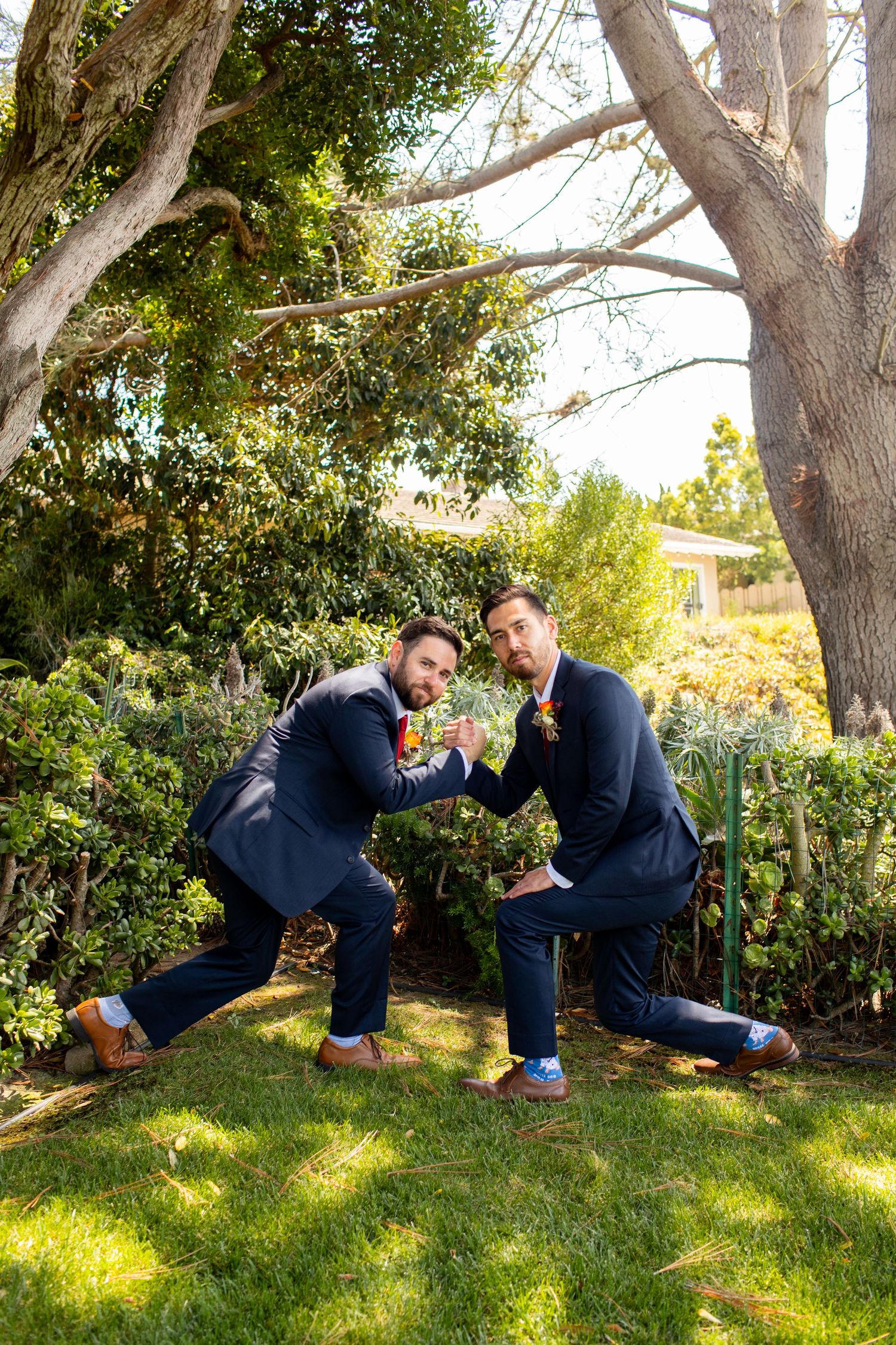 Two groomsmen in matching navy suits pose playfully on the lawn under large trees.
