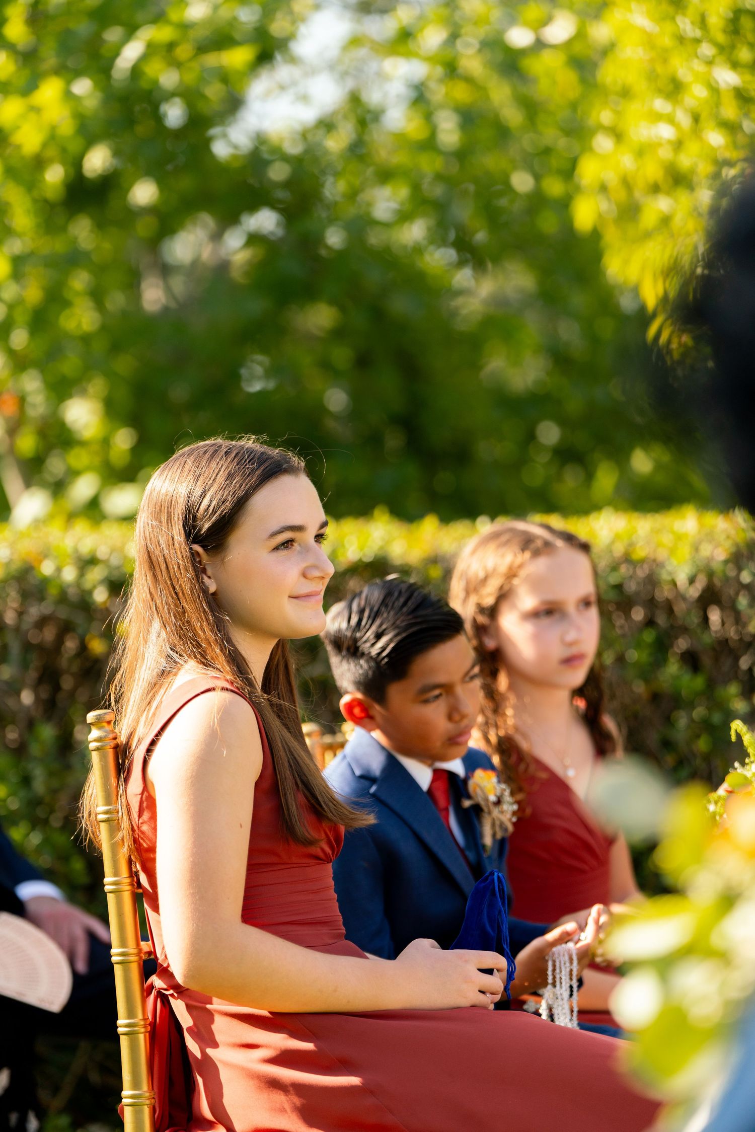 Wedding guests seated outdoors in formal attire at a garden ceremony with lush greenery in the background.