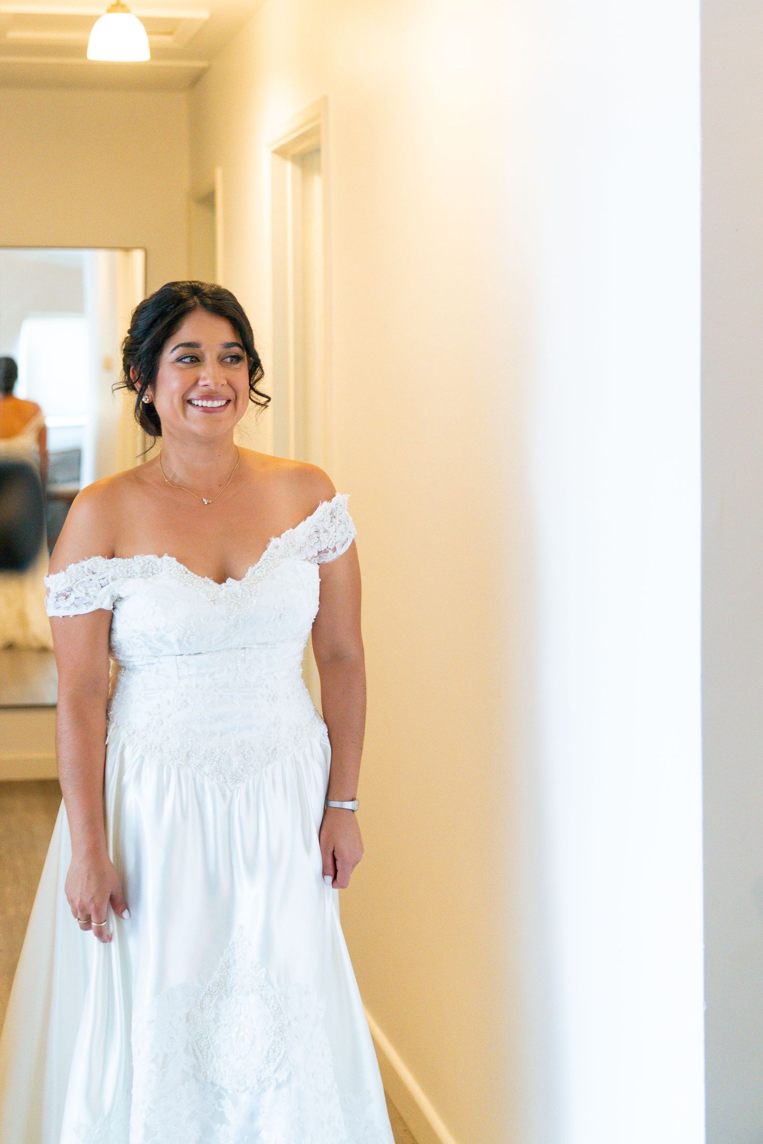 A radiant bride in an off-shoulder white gown stands in a sunlit hallway.