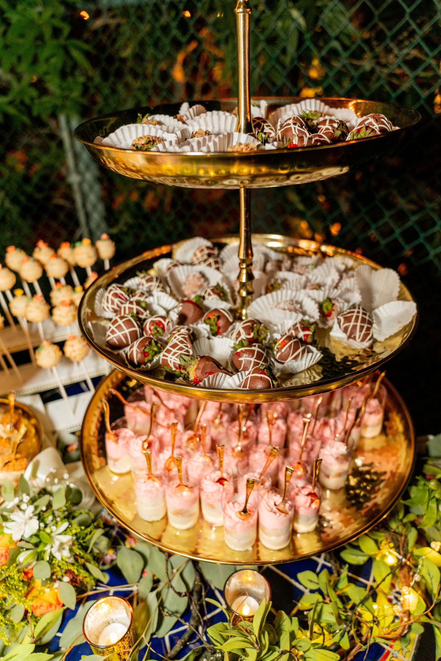 Elegant three-tiered dessert stand with chocolate covered strawberries and pink desserts at an outdoor reception.