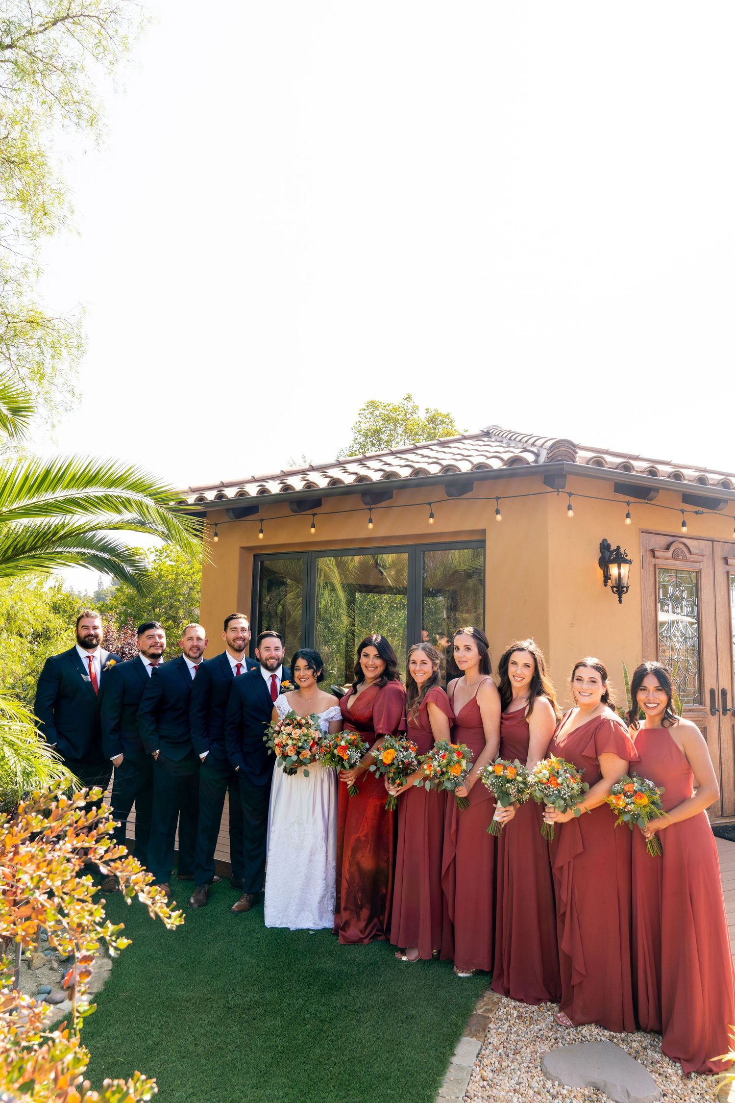 Wedding party in burgundy dresses and black suits poses together outside Spanish-style venue.