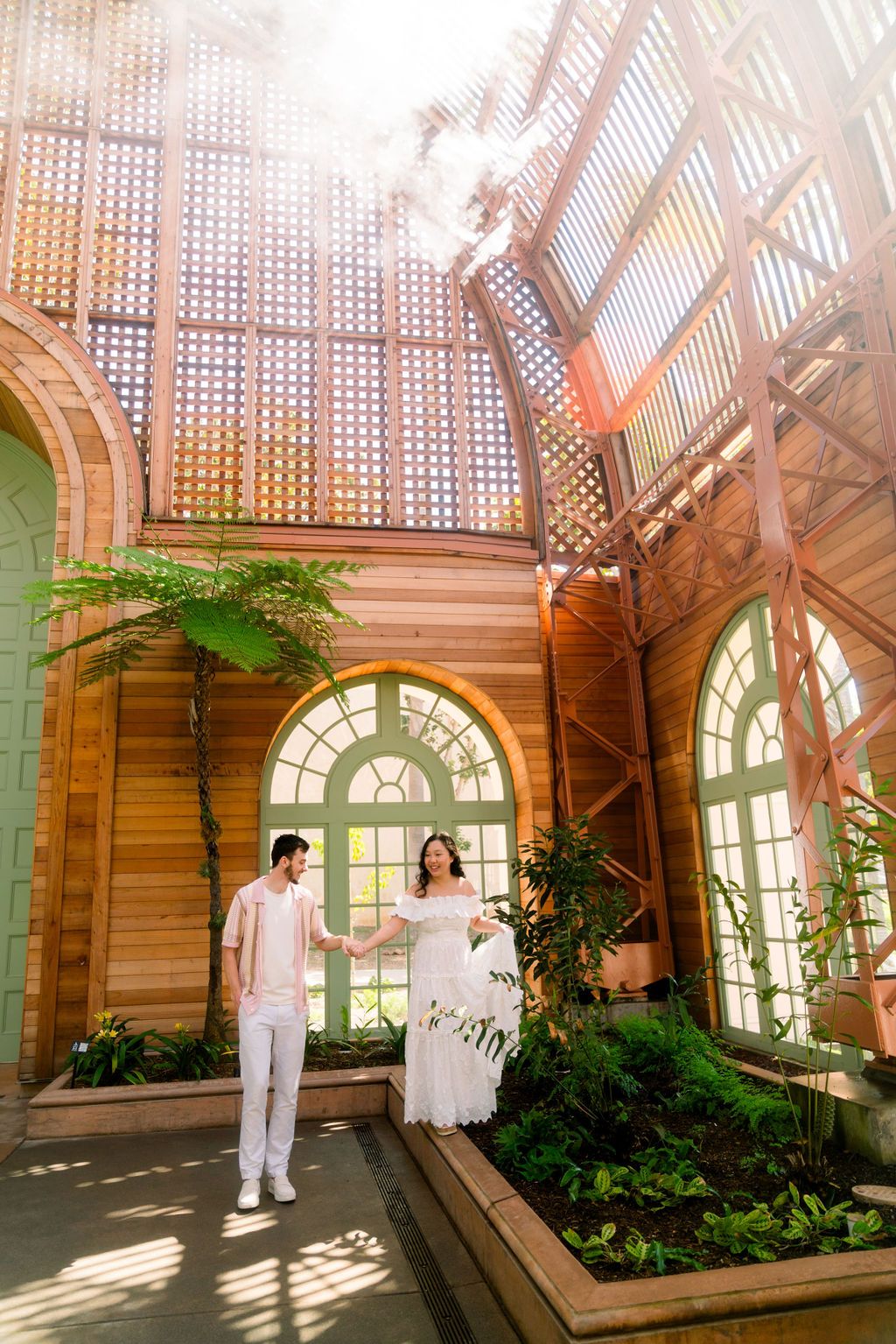 Elegant greenhouse wedding photo with sunlight streaming through glass ceiling panels and lush green plants surrounding the couple.