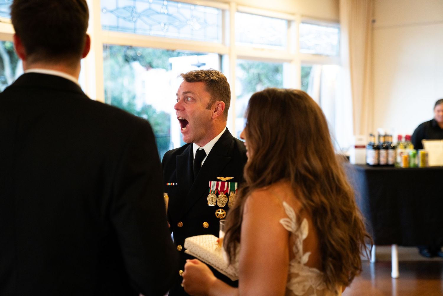 A naval officer in dress uniform laughs while socializing at an indoor event with guests and a bar in the background.