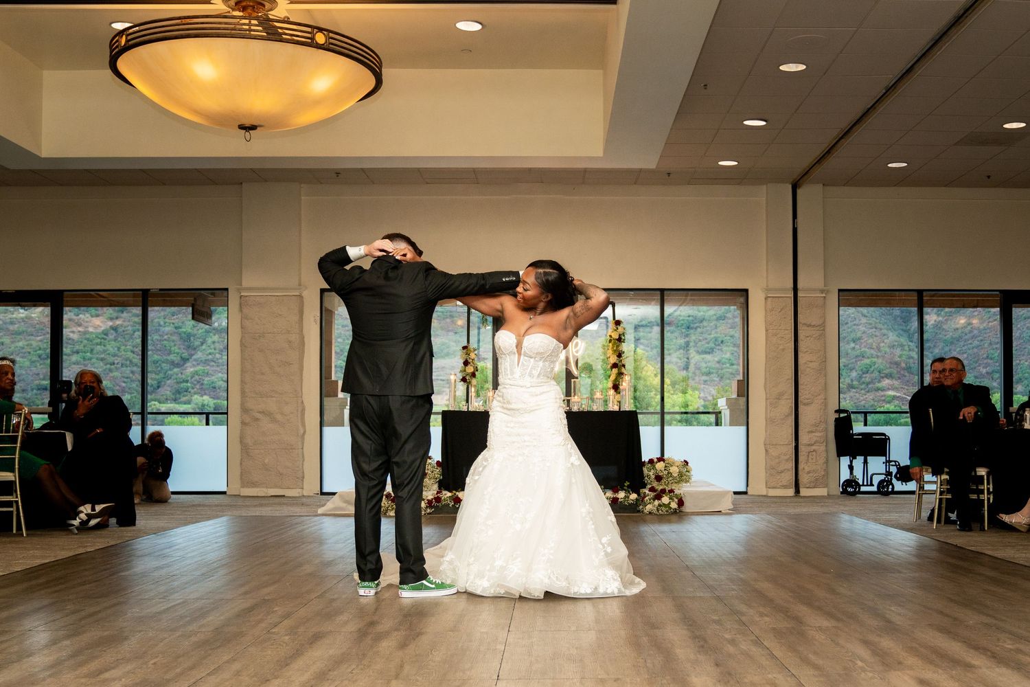 A couple performs a celebratory dance move in a reception hall with large windows.
