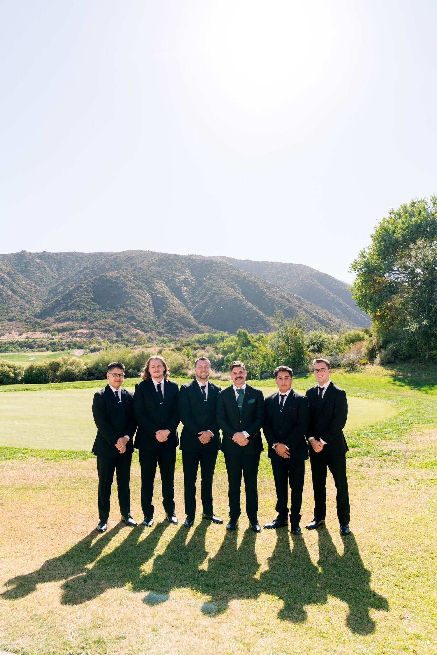 Wedding party in black suits standing in a line against mountainous backdrop.