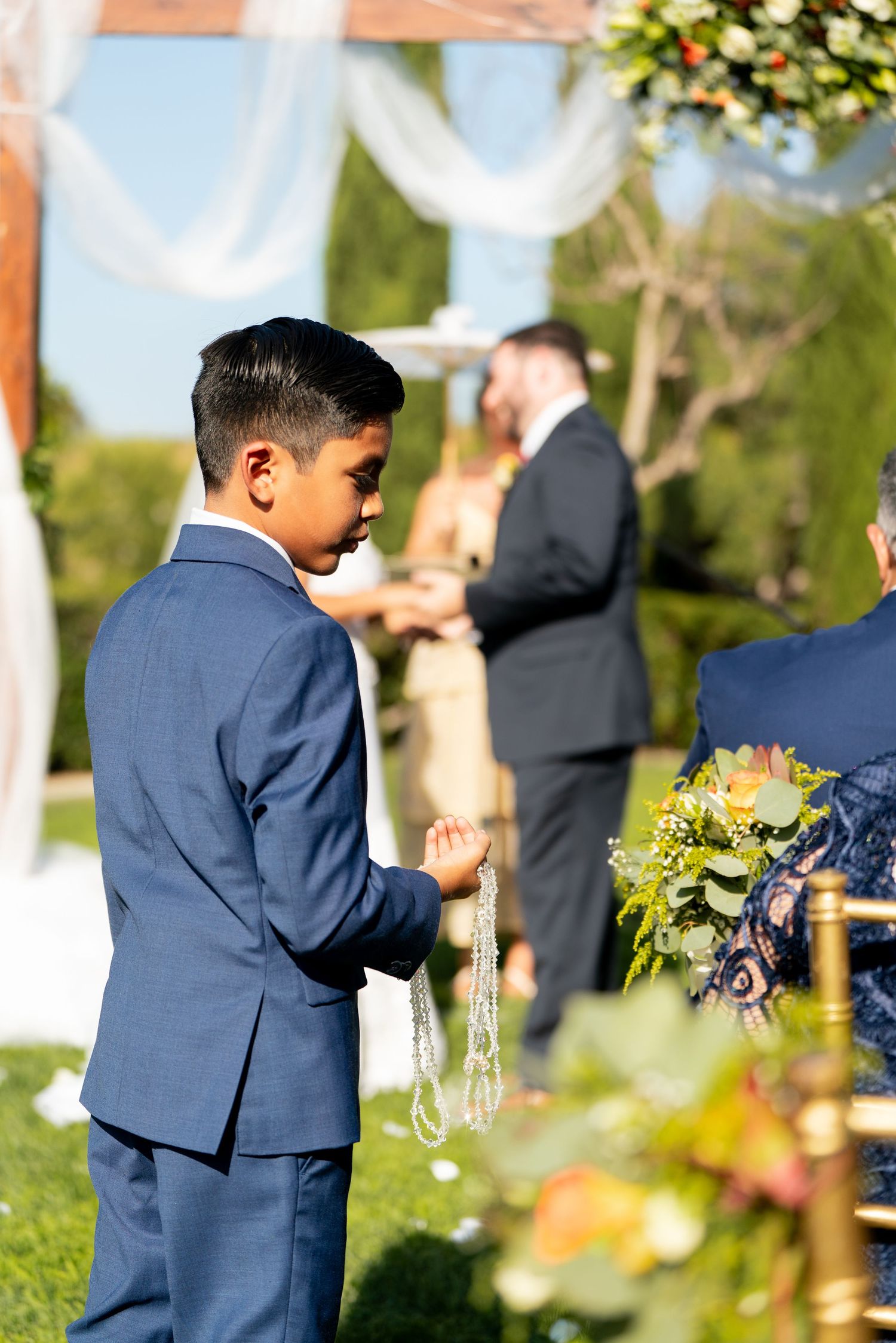 A wedding guest in a navy suit stands outdoors during a ceremony with white drapery and floral decorations.