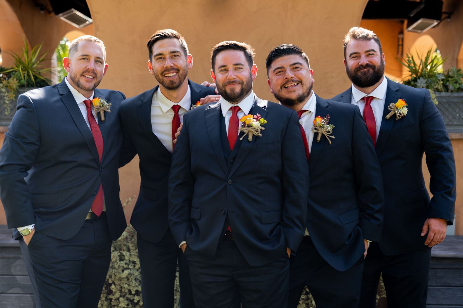 Groomsmen in dark suits with red ties pose together at an outdoor venue.