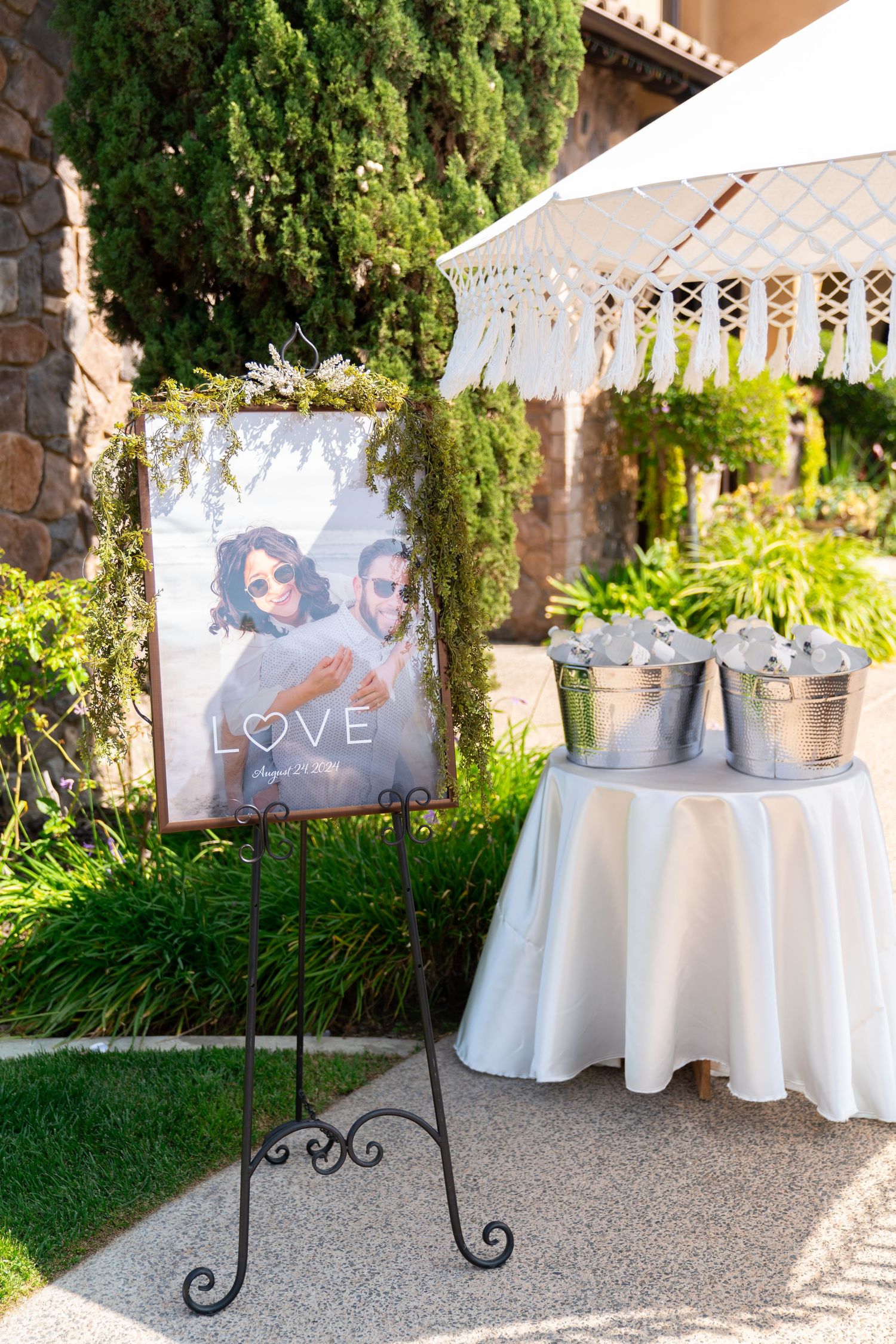 Wedding reception decor with a photo display stand and metal buckets on a white tablecloth in a garden setting.