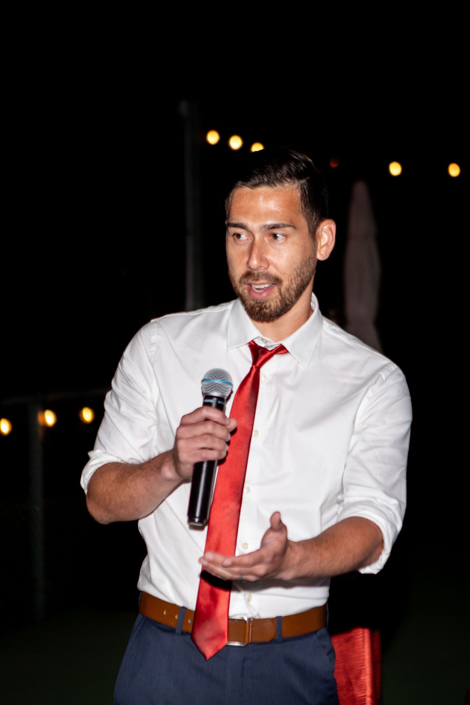 A speaker in white shirt and red tie addresses guests during an evening wedding reception.