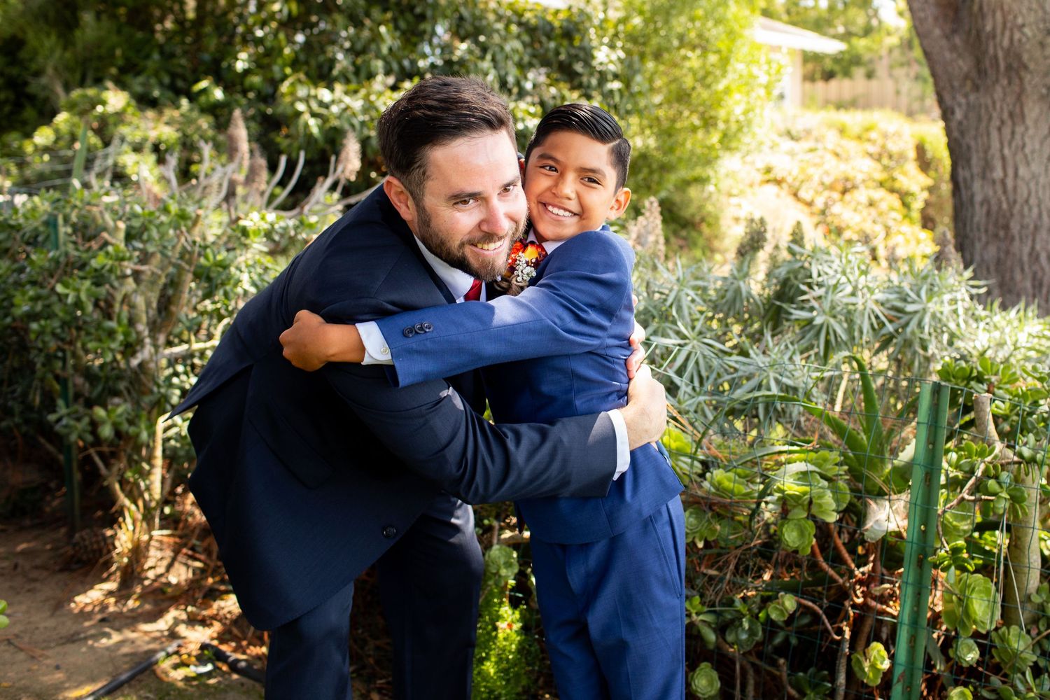 Two people in navy blue suits share a joyful embrace in a sunny garden setting.
