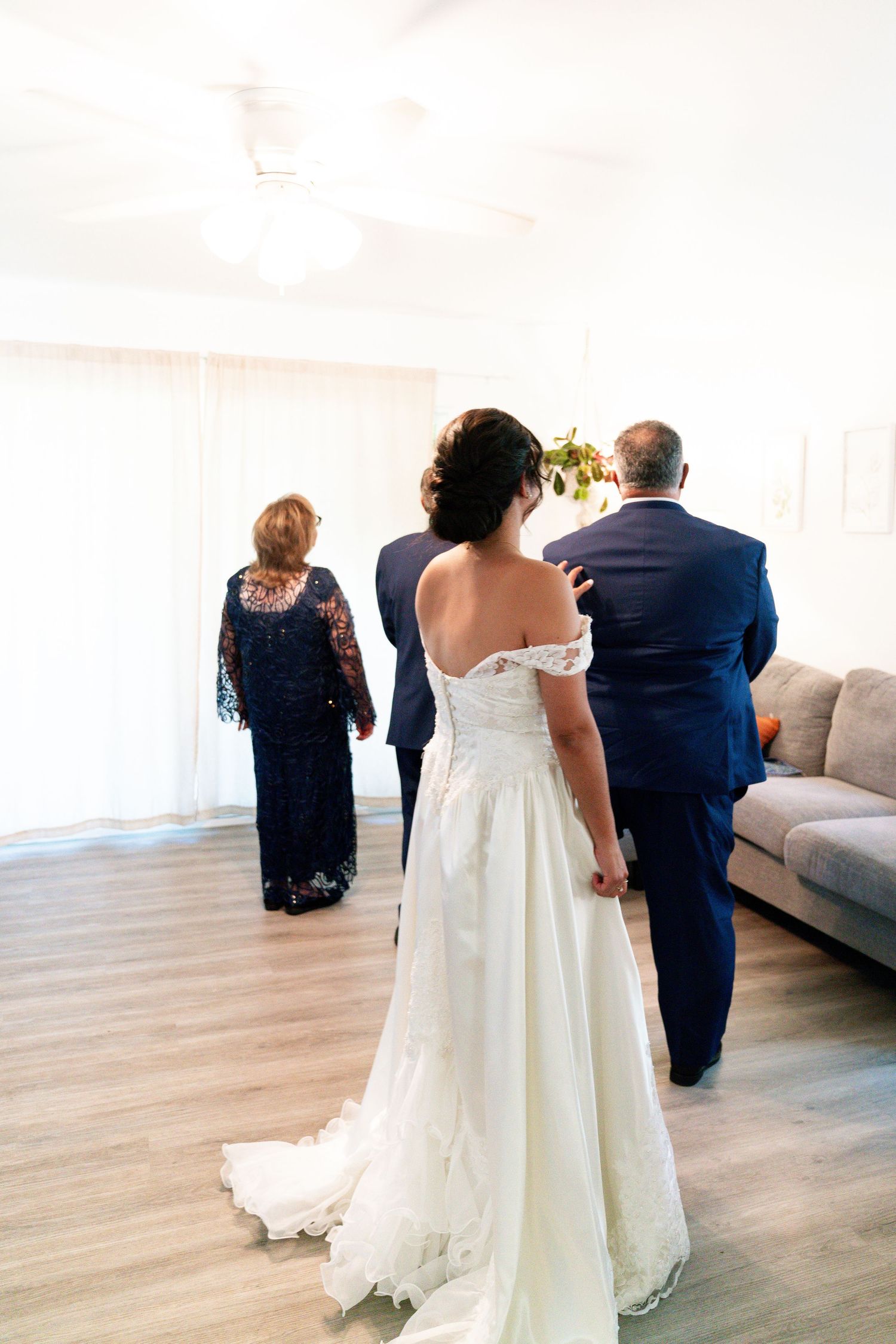 A bride in an elegant off-shoulder white wedding gown stands with wedding party members in a bright indoor setting.