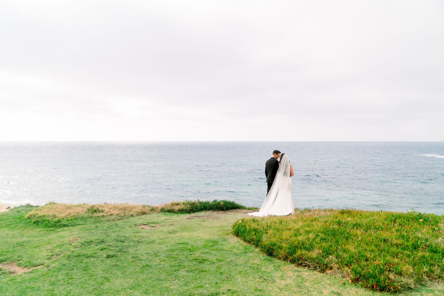 A couple stands on a green coastal cliff overlooking the ocean on their wedding day.