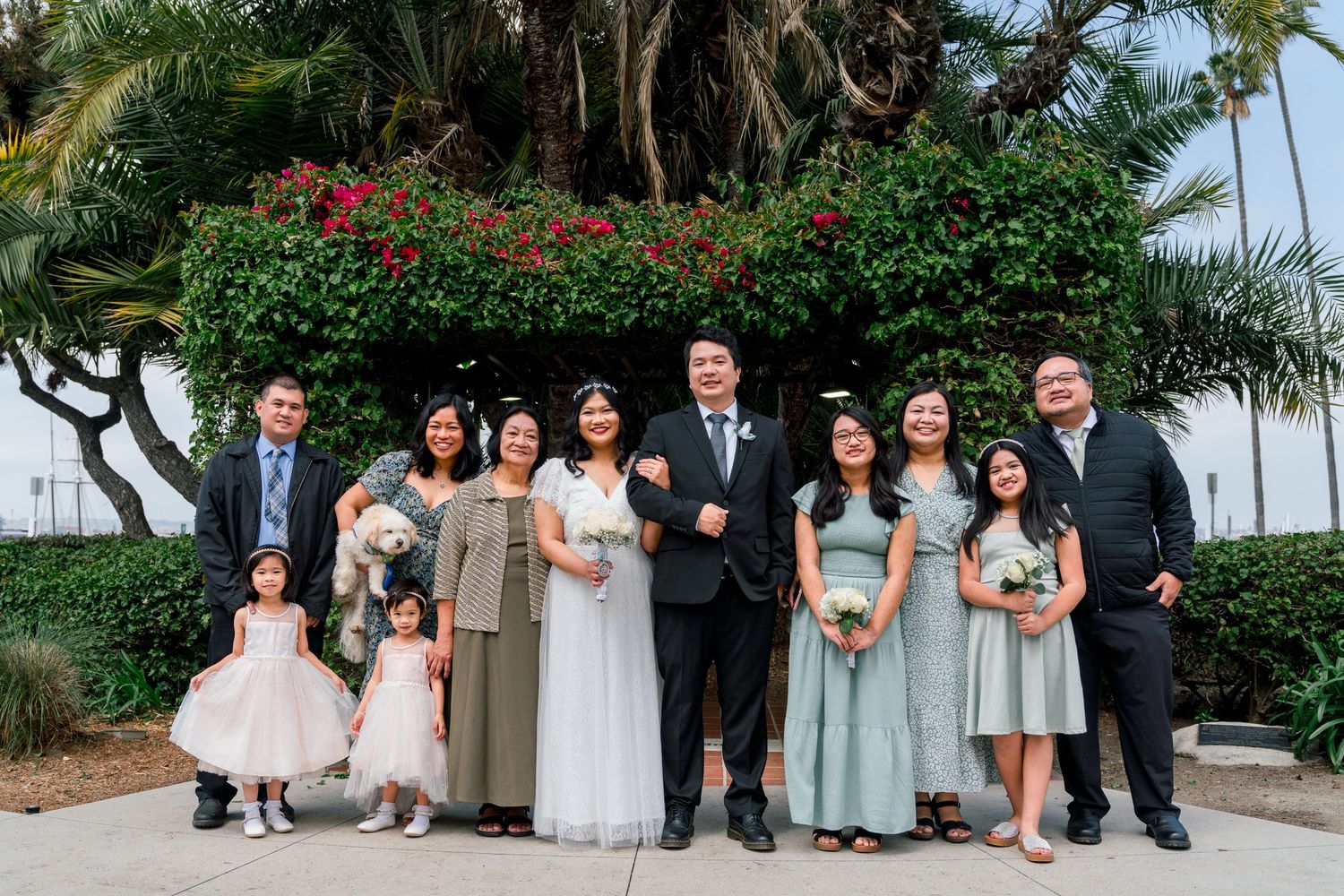 A wedding party stands together outdoors with palm trees and flowering bushes in a tropical garden setting.